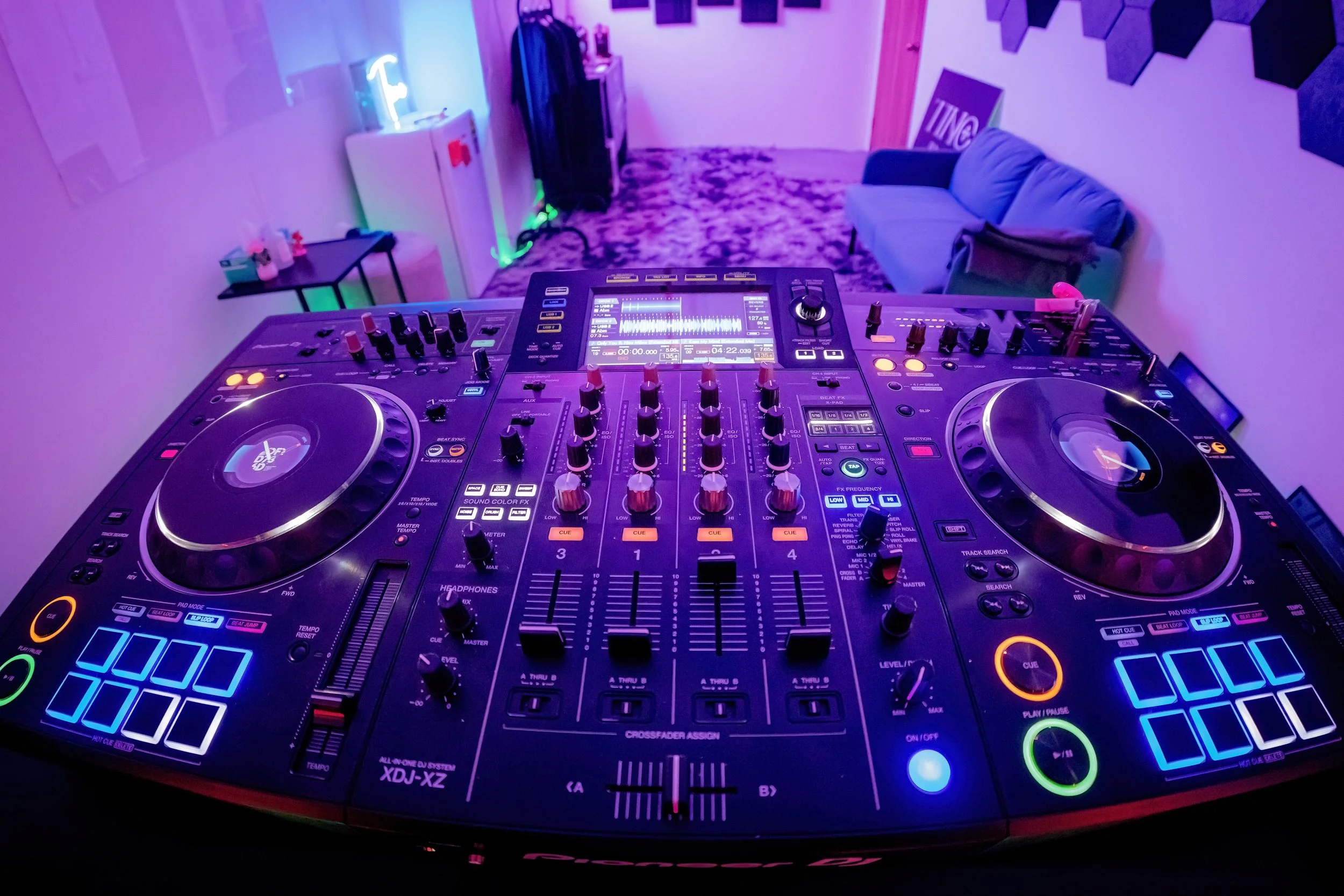 DJ turntables and mixer set up in a room with purple lighting, a sofa, and decorative wall art.