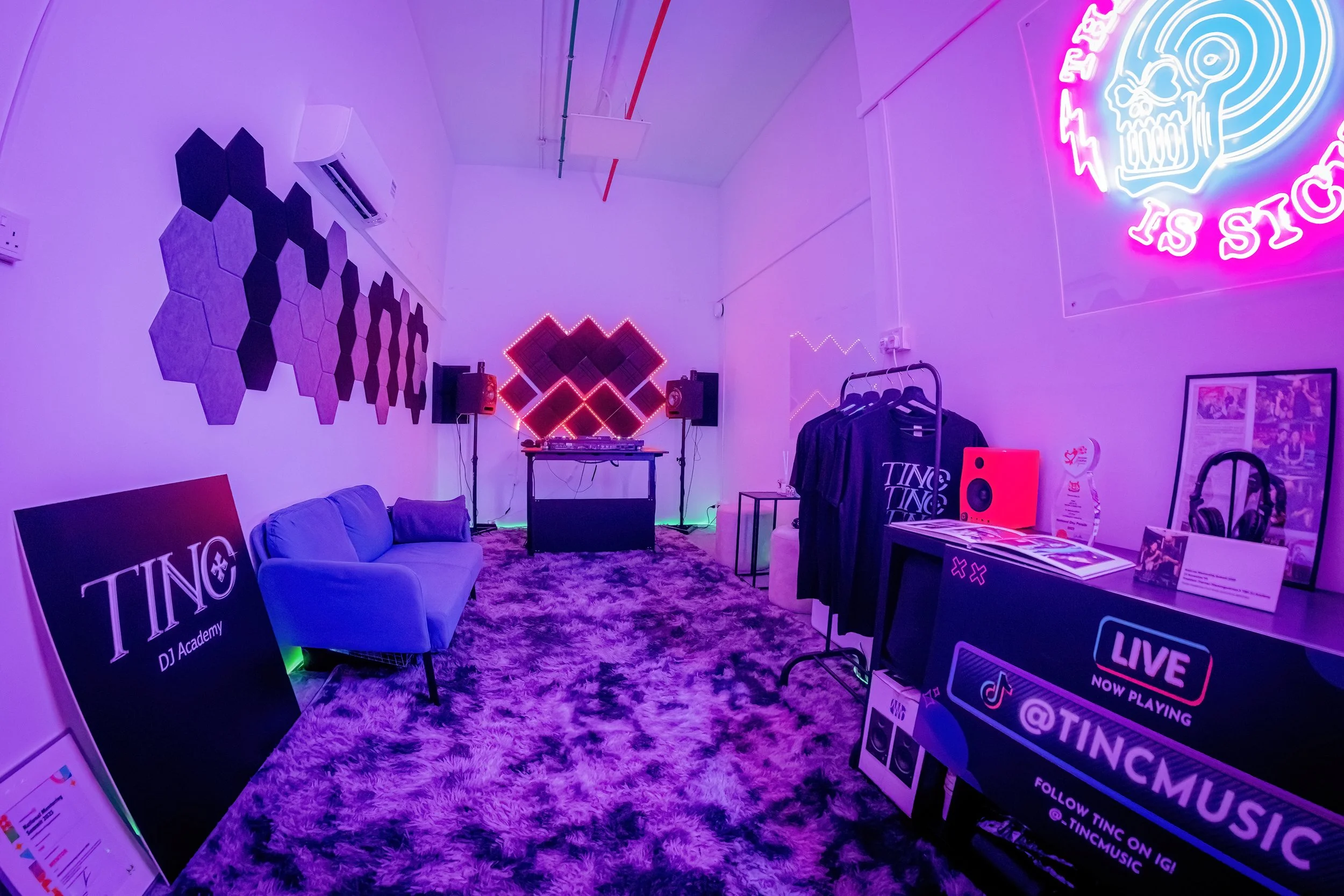 A room with neon lights, purple and pink hues, a DJ setup with speakers, a neon skull logo, a sign that says 'TINC DJ Academy', a couch, a clothing rack, and music-related decorations.