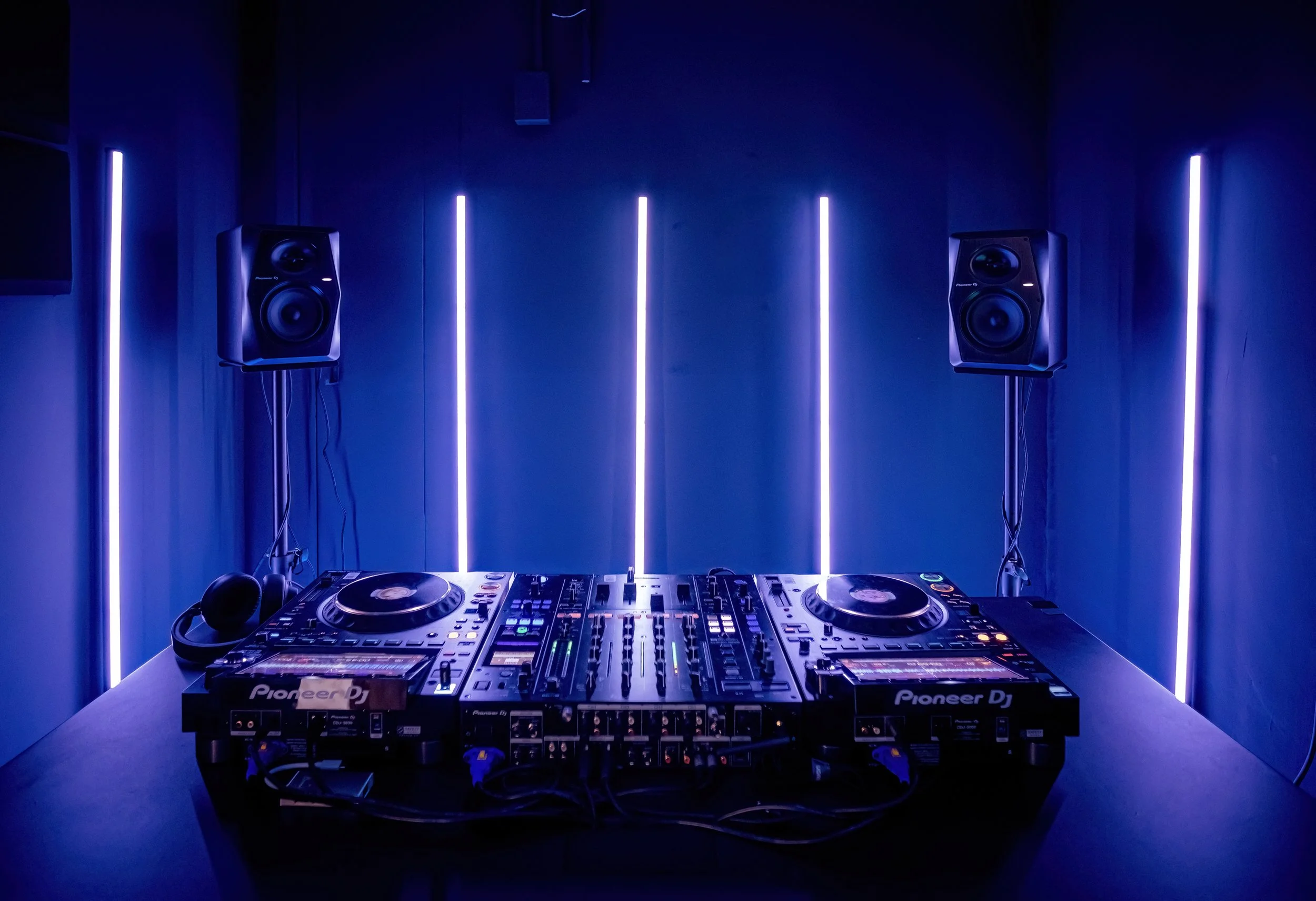 DJ mixing console with two Pioneer DJ turntables set up in a dark room with blue LED vertical lights on the wall behind.
