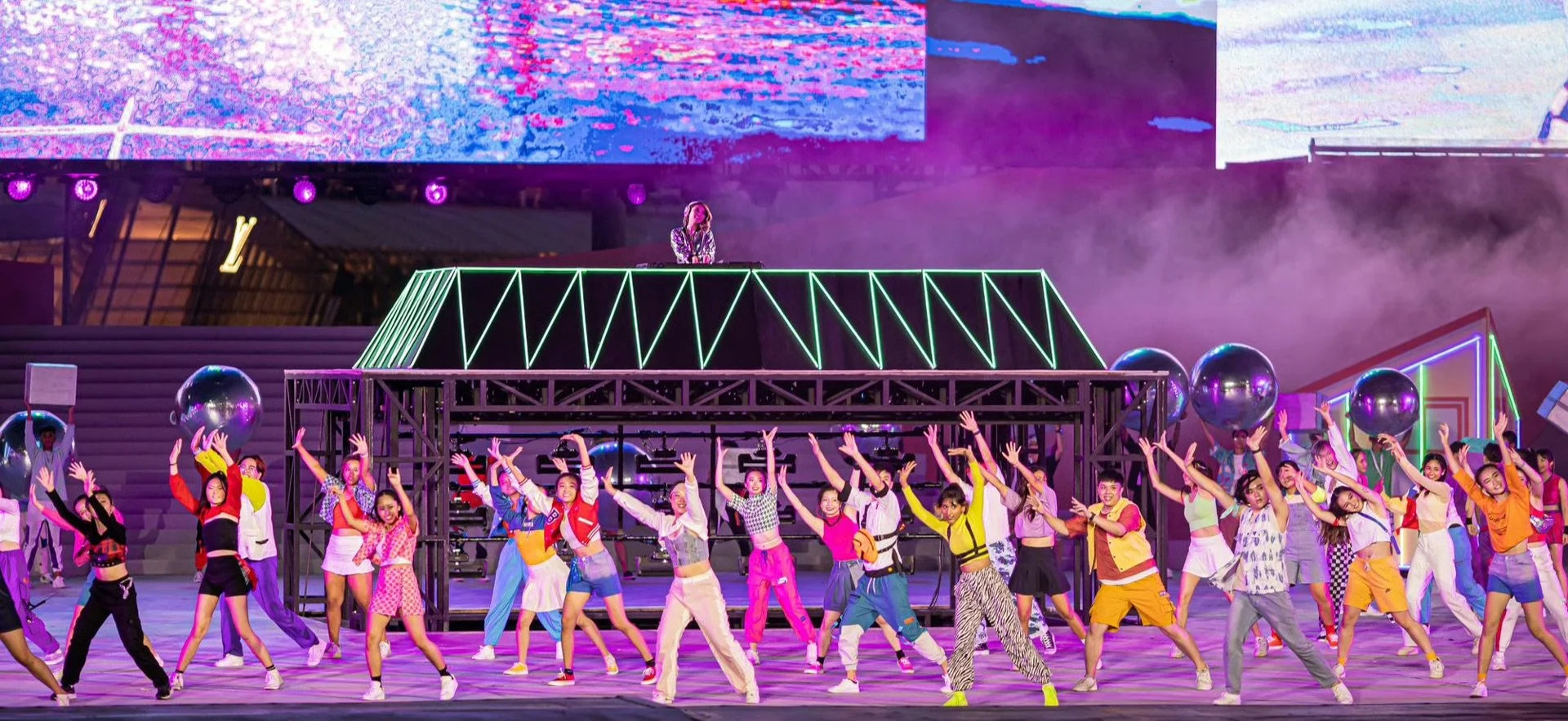 A large group of young performers dancing on stage with vibrant, colorful lighting, neon props, and a DJ on an elevated platform in the background at an outdoor event.