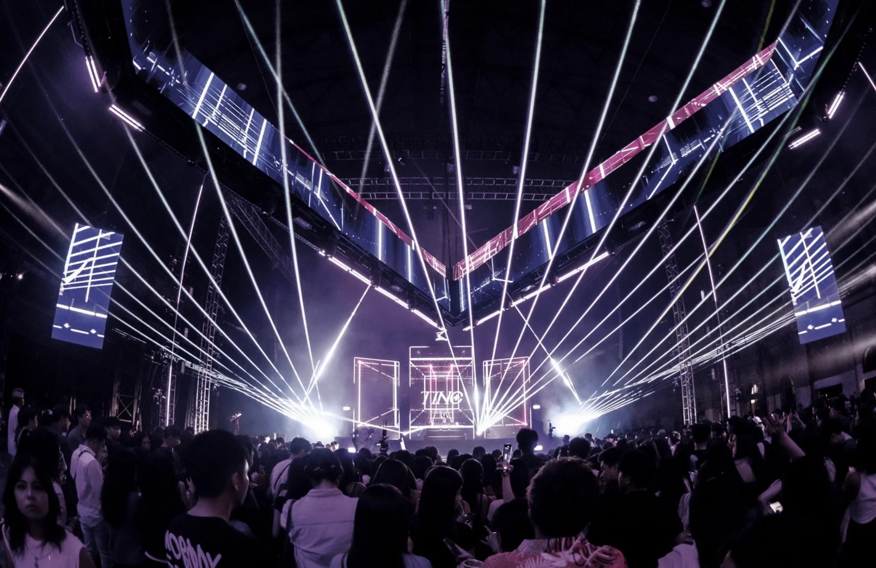 A large indoor concert or event with a stage and a crowd of people. The stage has bright laser lights and screens displaying visual graphics.