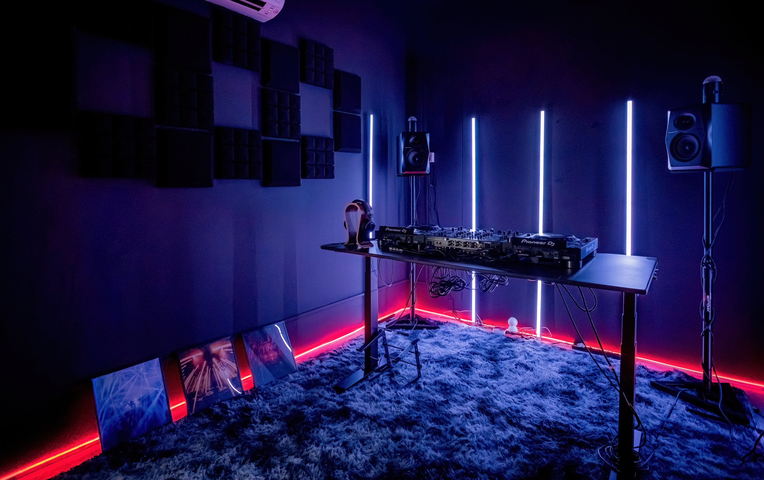 A dimly lit music studio with neon blue and red lighting, a DJ setup with turntables, and black acoustic foam panels on the wall.
