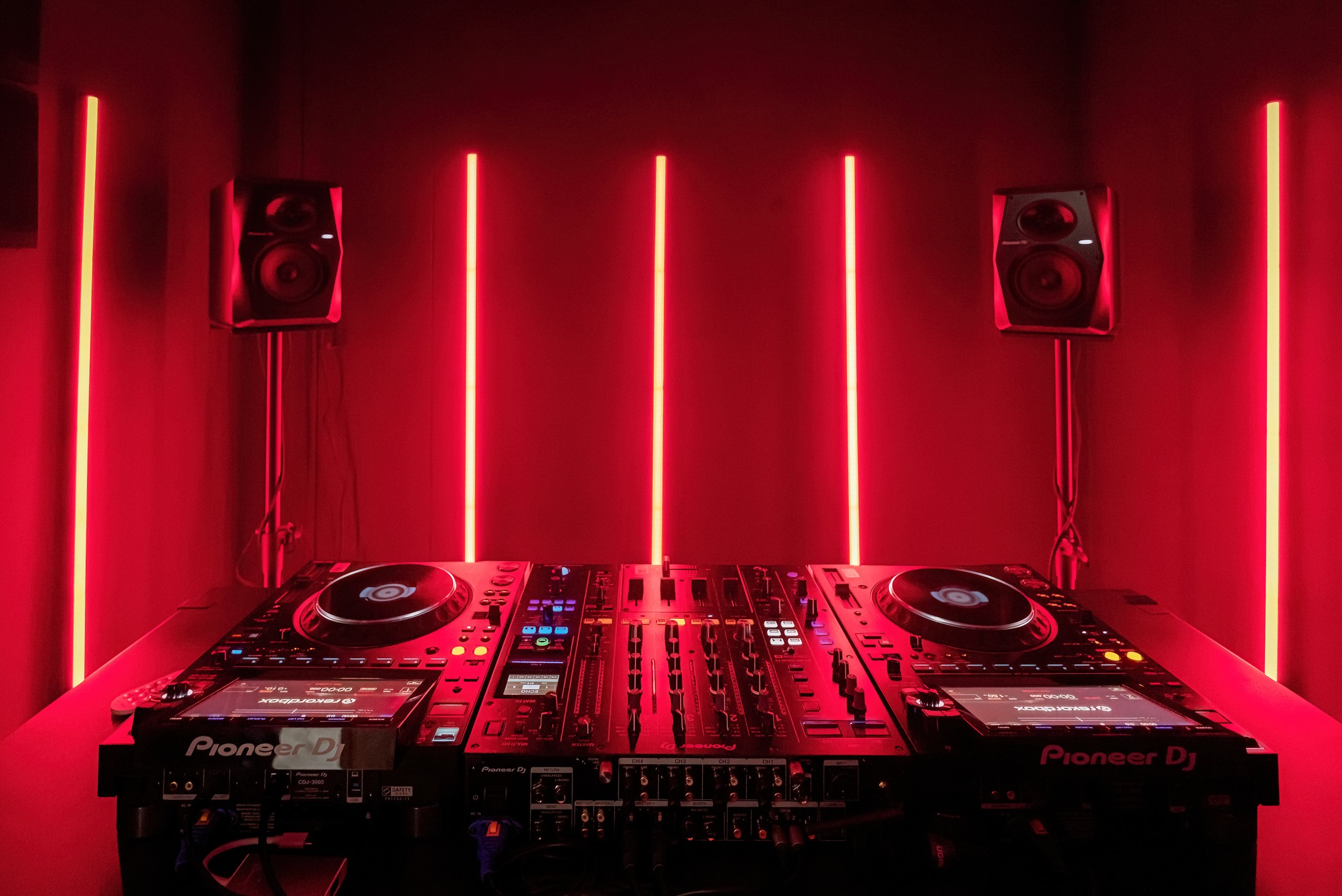 DJ setup with Pioneer CDJ turntables, a mixer, and two speakers in a red-lit room.