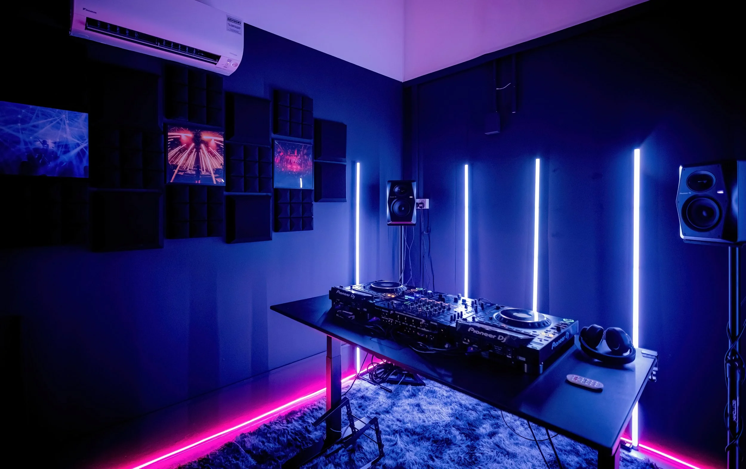 A modern DJ setup with Pioneer DJ equipment on a black table, surrounded by black and white speakers, featuring vibrant purple and pink LED lighting, and wall-mounted screens displaying digital visuals in a dark room.