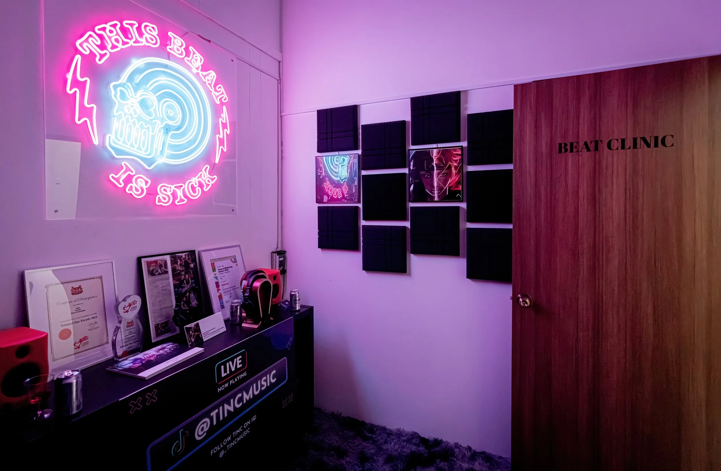 Interior of a music studio with a neon sign that says 'This Beat Is Sick' and a wall decorated with black acoustic panels and a neon portrait of a woman. A table with awards, framed certificates, headphones, and drinks is visible, along with a door l