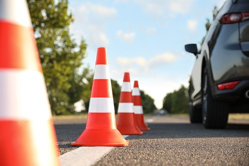 Traffic cones on the side of the road near parked cars, with trees and a blue sky in the background.