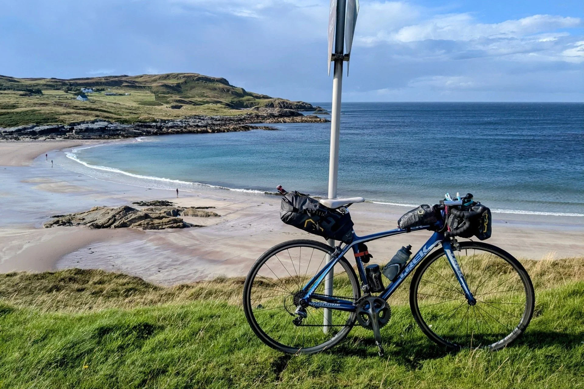 A blue touring bicycle with accessories leaning against a metal pole on a grassy hilltop overlooking a sandy cove with shallow water, rocks, and green hills in the distance.