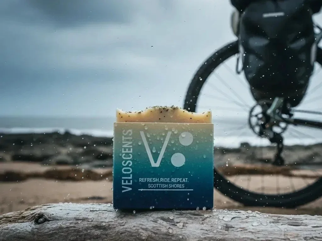 A bar of Veloscents soap with a blue and white label, placed on a rock near the shoreline with a bicycle in the background, under cloudy skies.