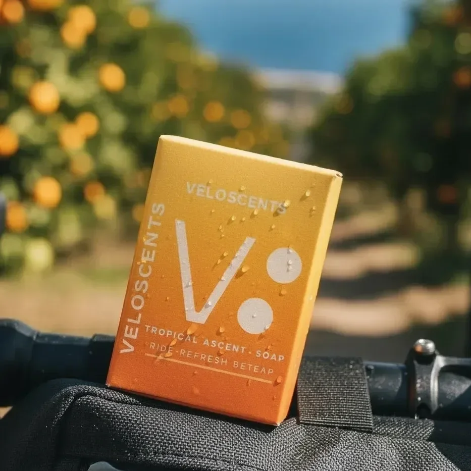 A yellow-orange box of Velosent tropical ascent soap with droplets of water on it, resting on a black surface outdoors with blurred green foliage and orange flowers in the background.
