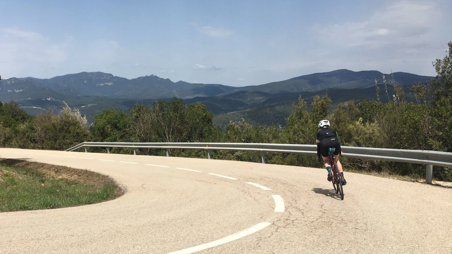 Cyclist riding on a winding mountain road with lush green trees and mountains in the background.