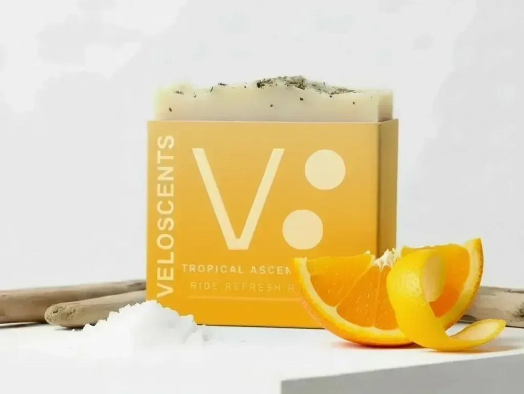 A yellow box labeled 'Veloscents' with the words 'Tropical Ascent' and 'Ride Refresh' underneath, containing a soap bar topped with black specks, an orange wedge, and a lemon peel.