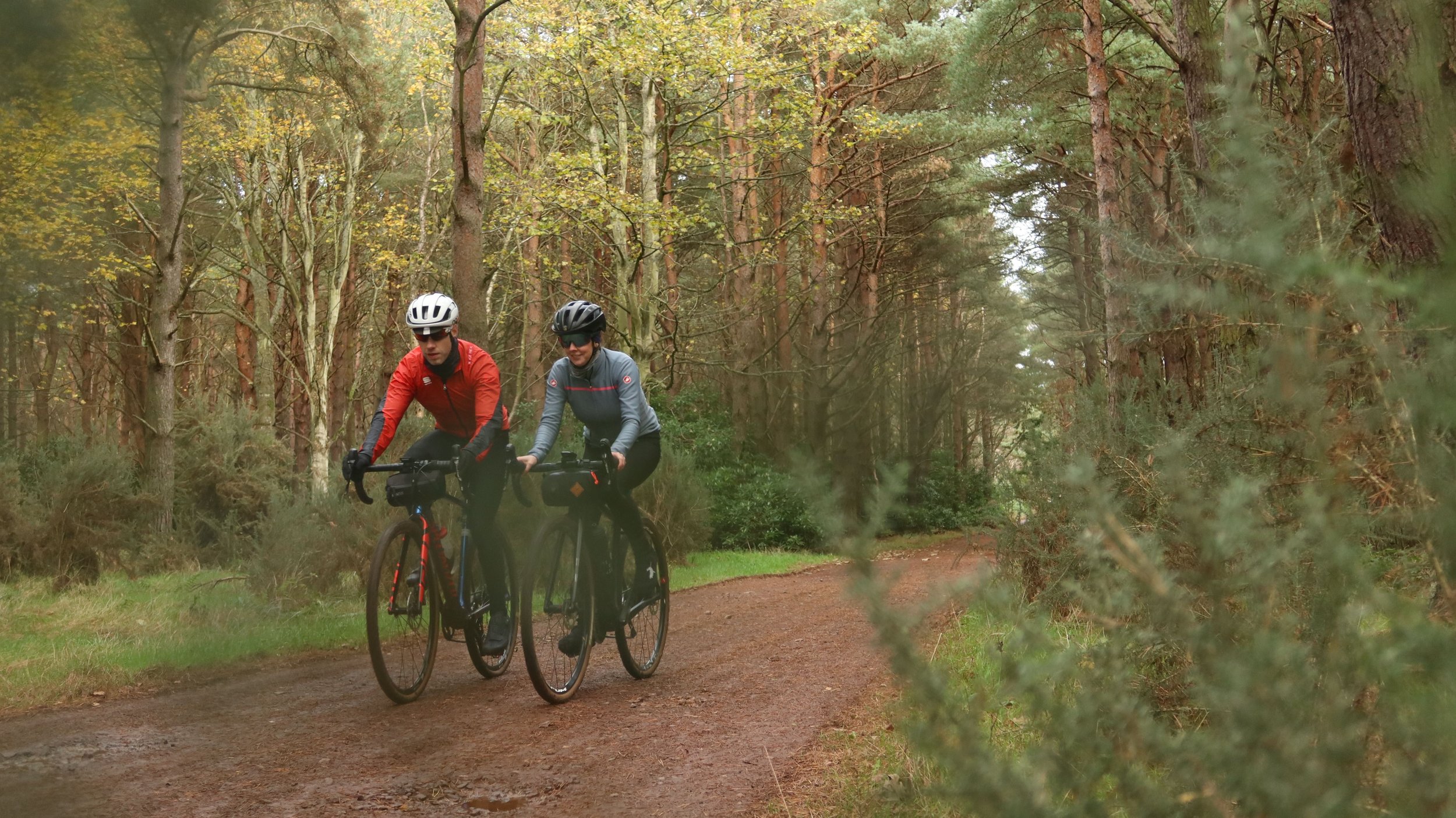 Two cyclists riding on a dirt trail through a forest with tall trees and green foliage.