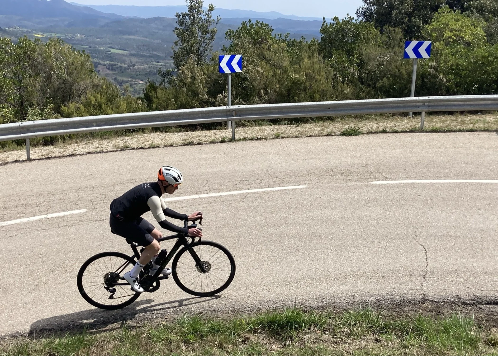 A person riding a black bicycle along a curved mountain road with a metal guardrail. The cyclist is wearing a white helmet, black cycling clothes, and white socks. The background features trees, green hills, and mountains under a partly cloudy sky.