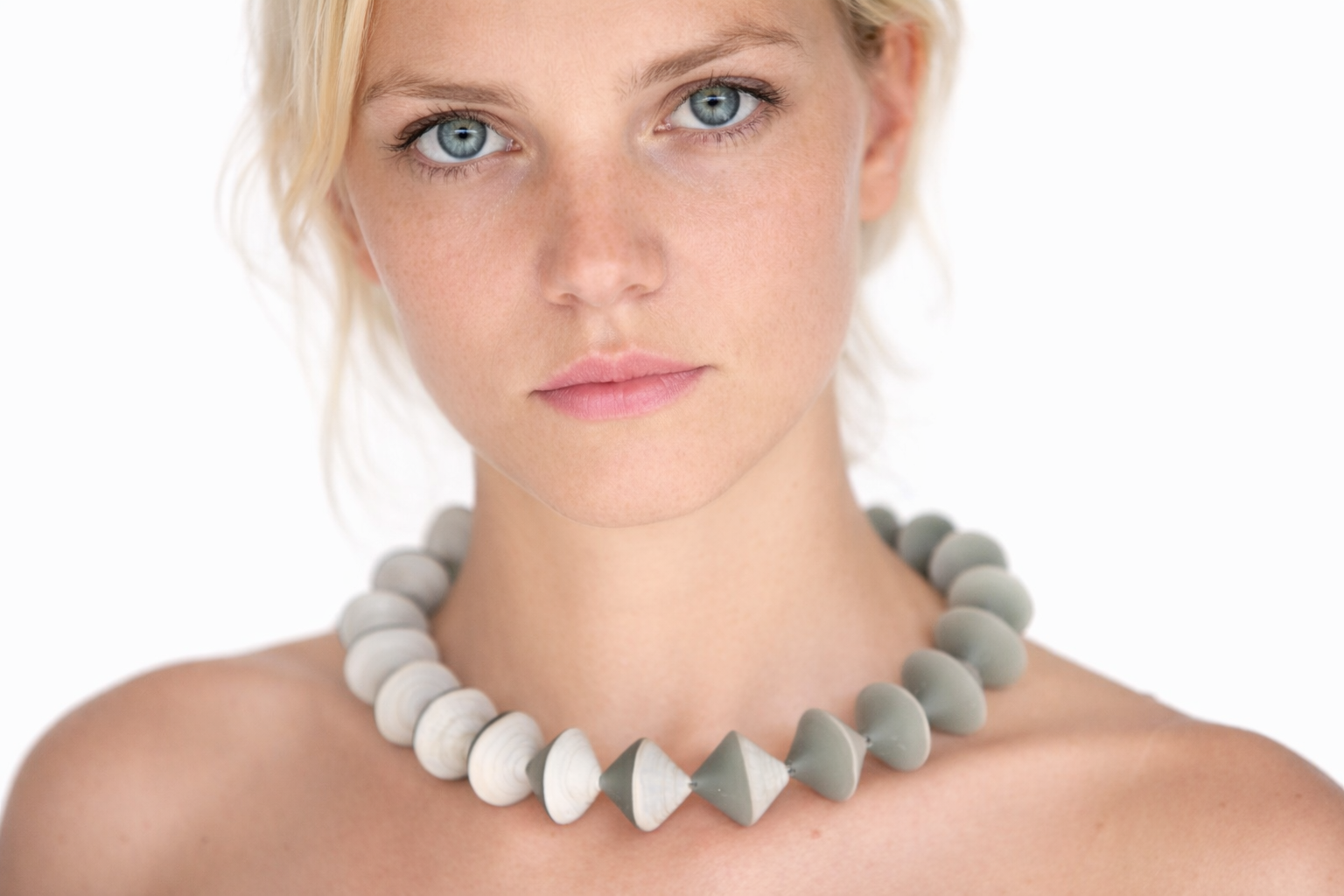 Close-up of a young woman with blue eyes, fair complexion, and short blonde hair, wearing a chunky white and gray beaded necklace.