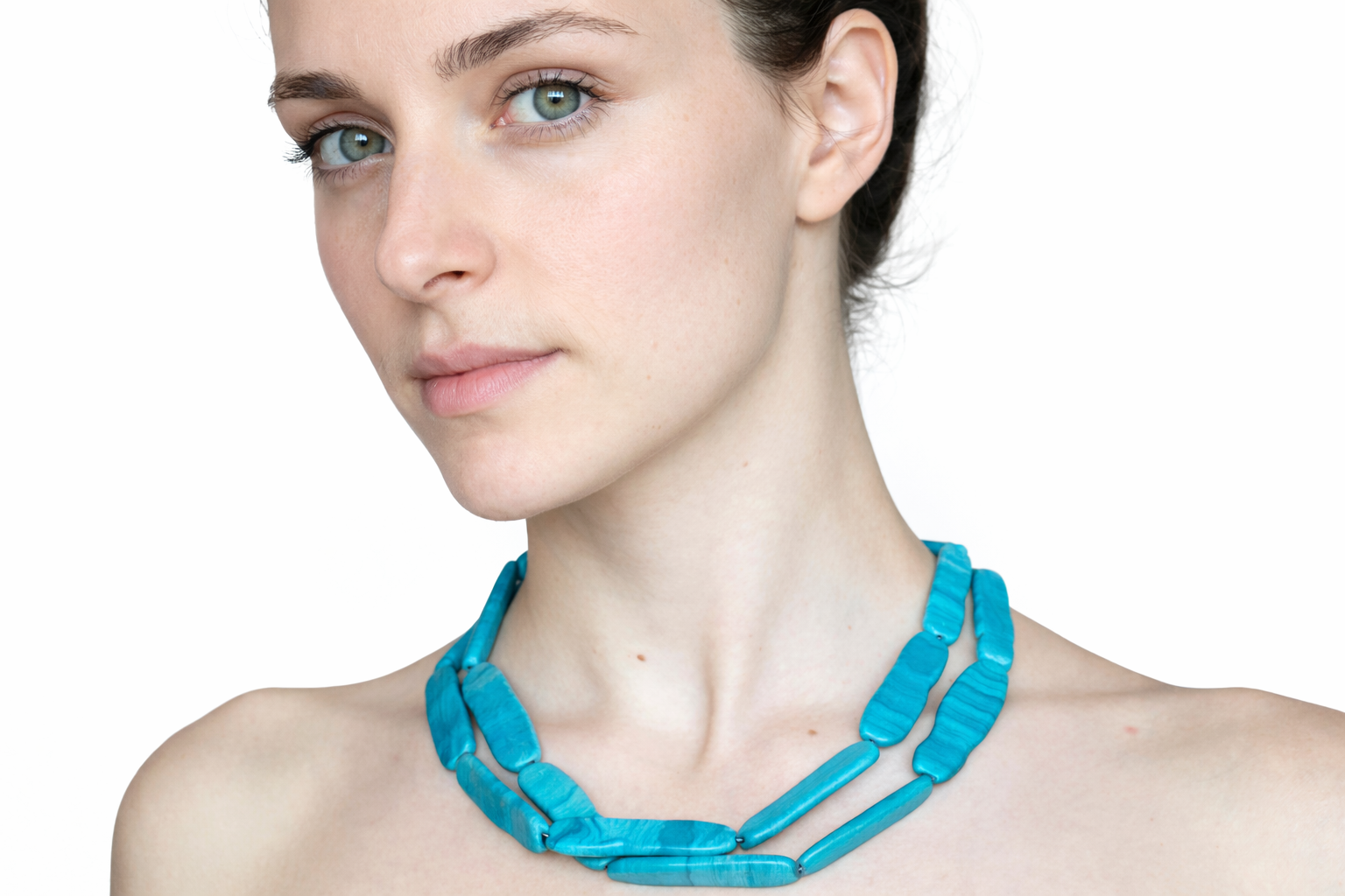 A close-up portrait of a young woman with fair skin and blue eyes, wearing a chunky turquoise necklace, with a white background.