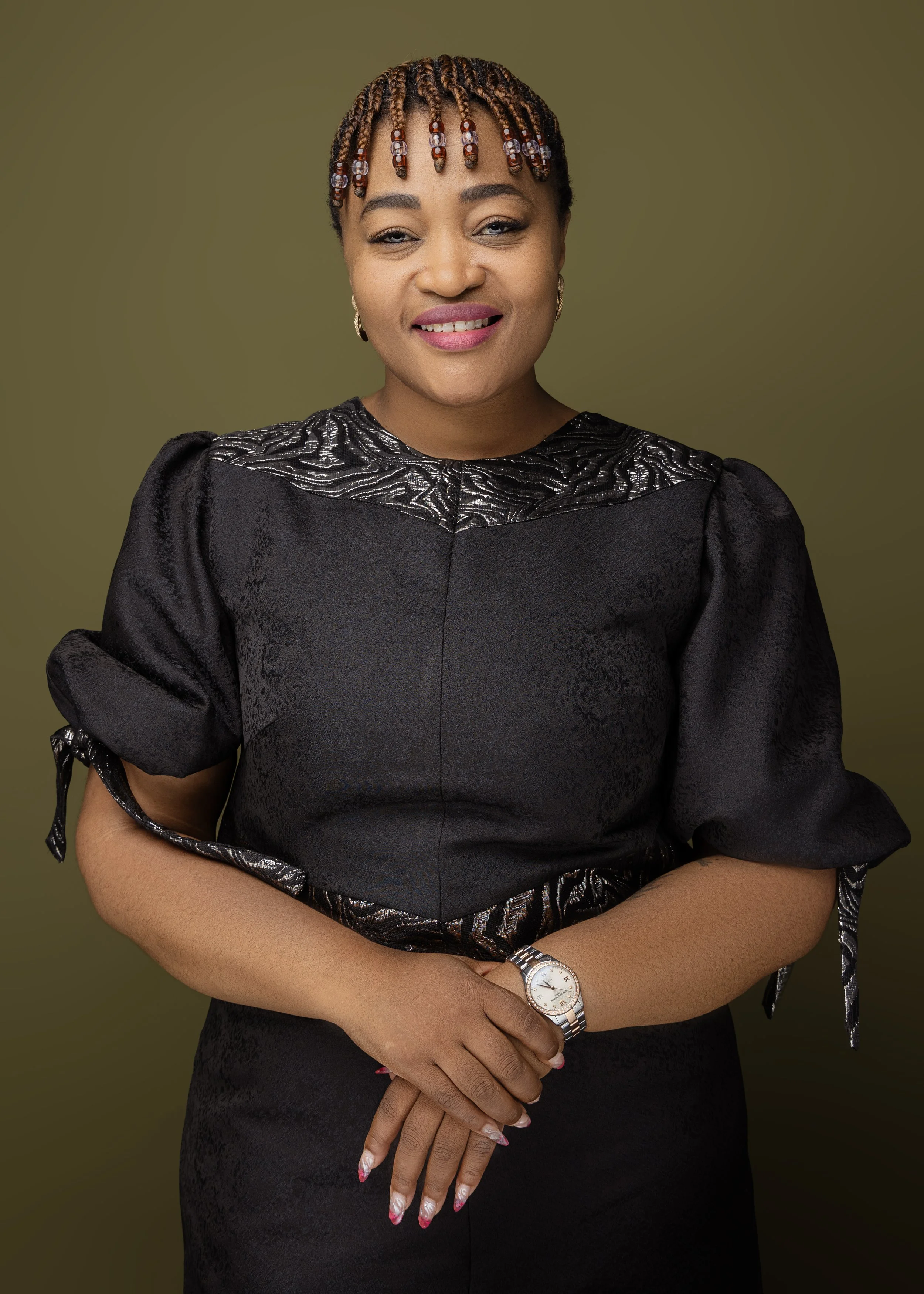 A woman with braided hair and beads, wearing a black dress with lace details and puffy sleeves, standing against a plain background.