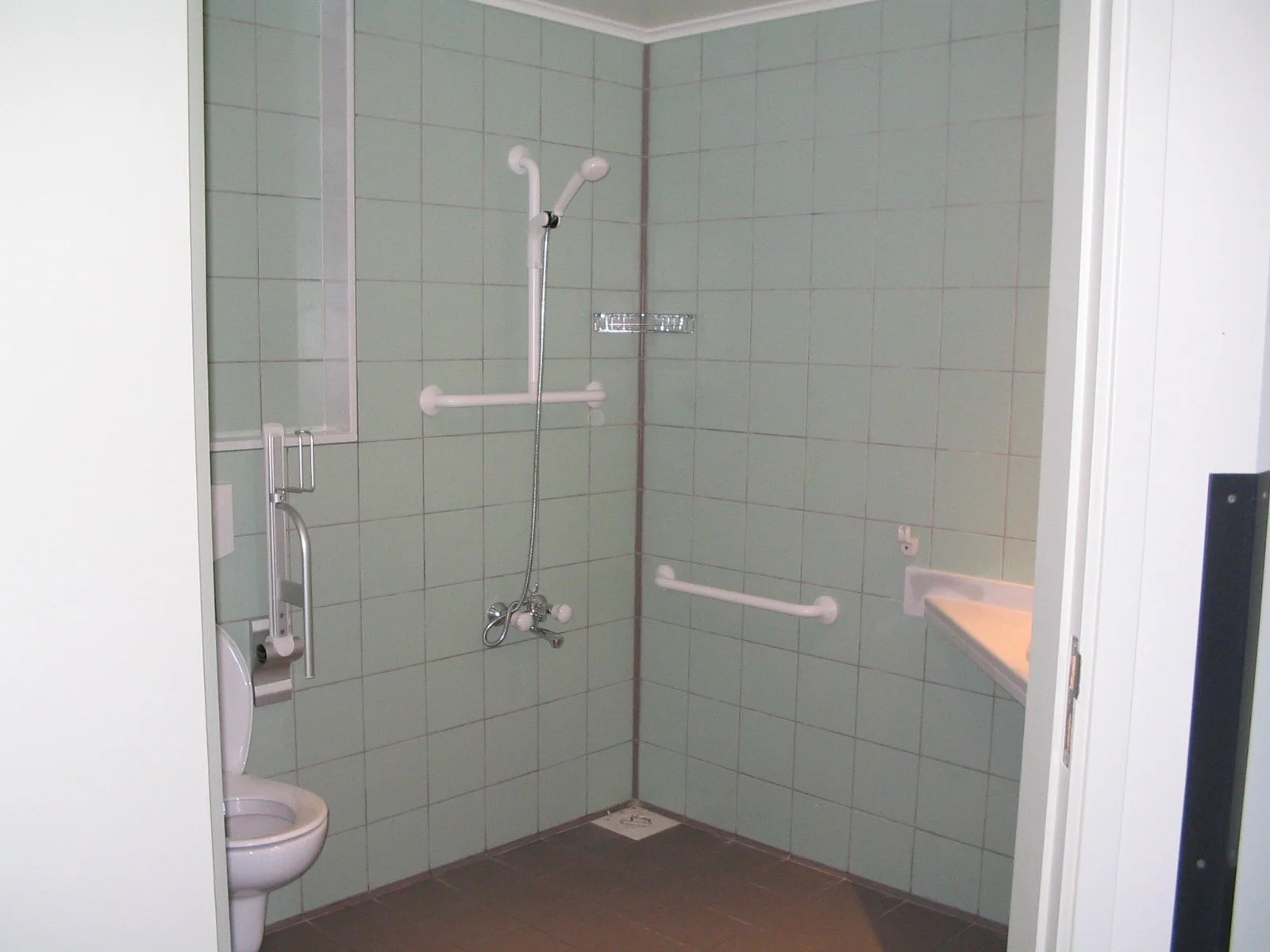 Accessibility shower with a fold-down seat, grab bars, handheld showerhead, mirror, and a small sink in a tiled bathroom.