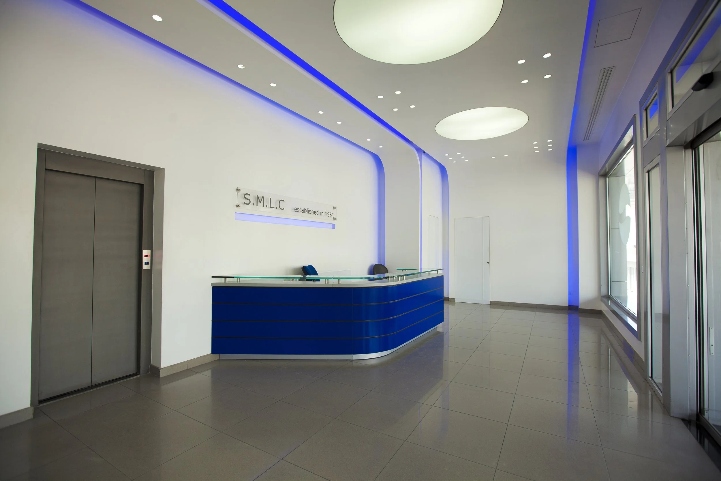 Modern lobby with a reception desk, an elevator, and large glass windows with automatic doors, featuring a sleek white and blue color scheme with LED lighting.