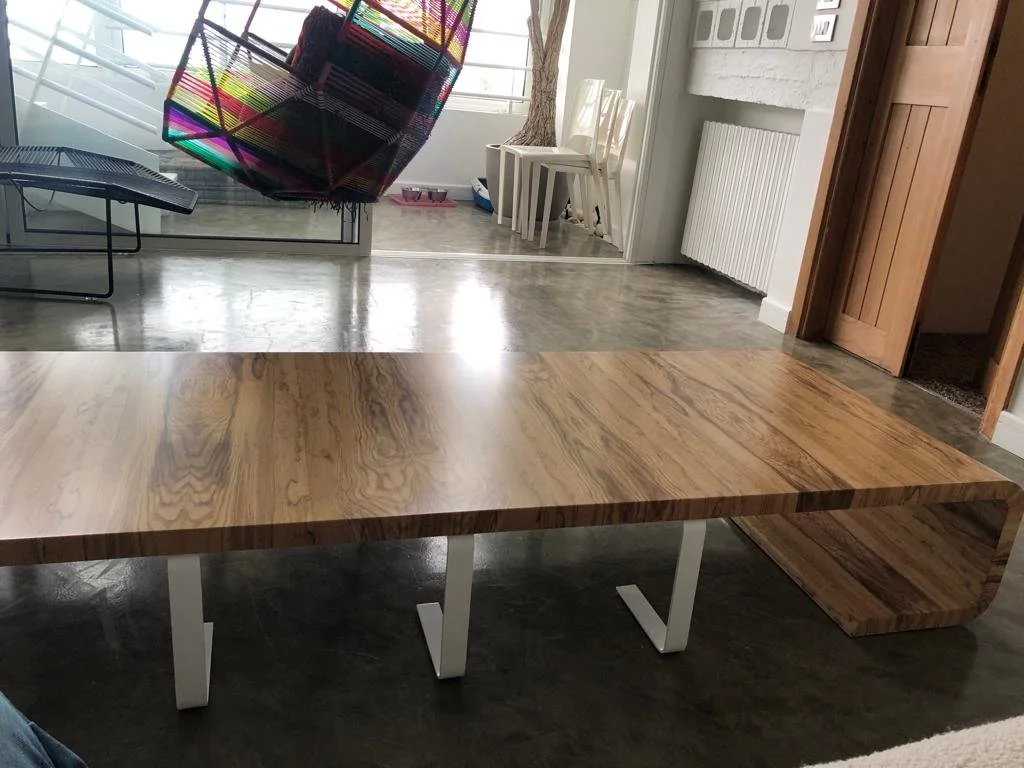 A wooden table with a light finish and white support legs, positioned on a dark floor in a modern living space.