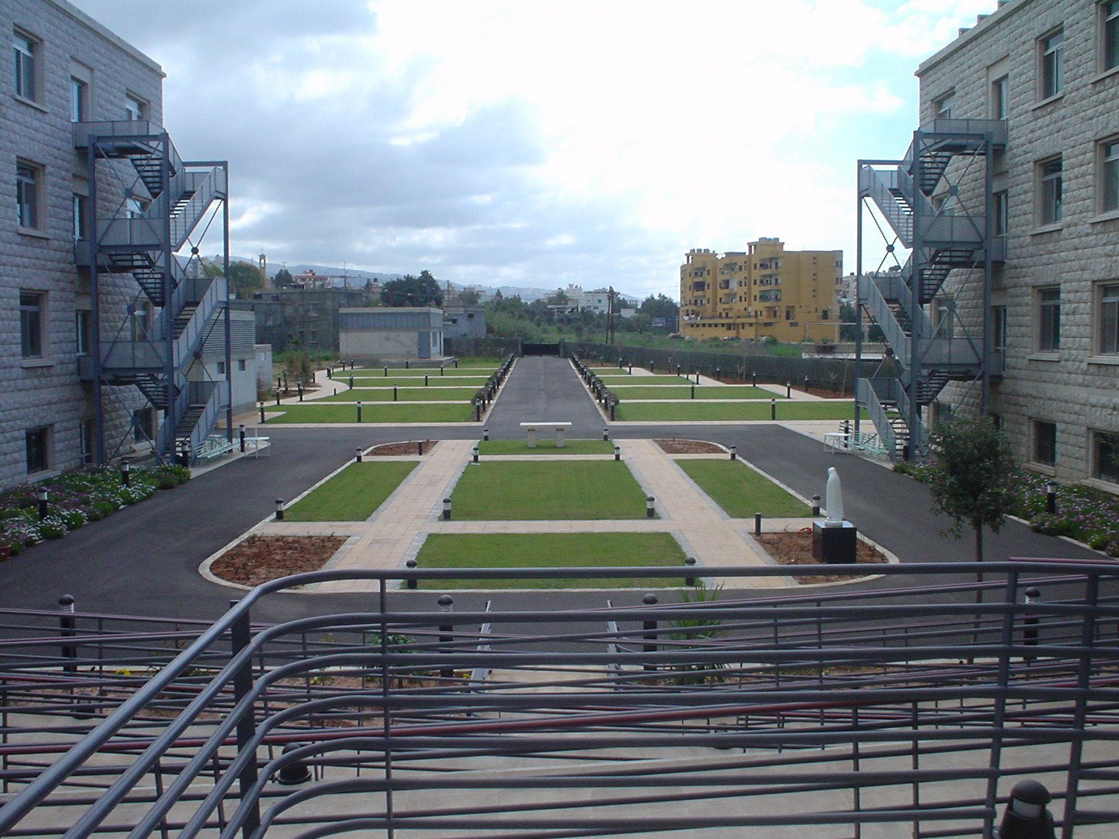 View of a courtyard with green grass, paved walkways, outdoor benches, and small trees. Surrounding buildings have gray stone facades and fire escape stairs. The sky is cloudy.