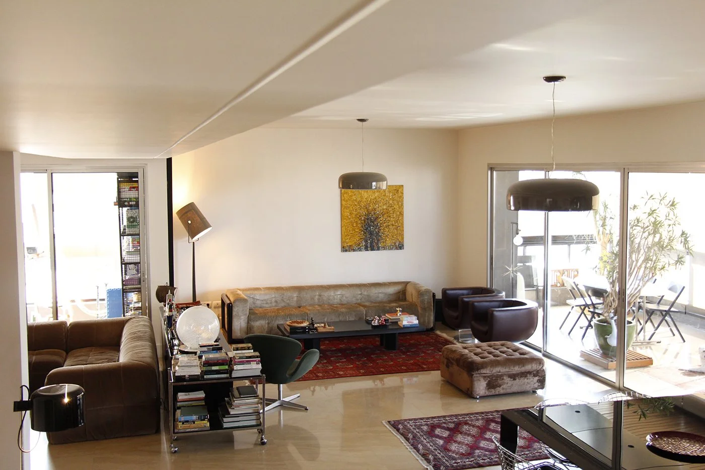 Living room with brown leather sofas, a glass coffee table, a colorful rug, and a sliding glass door leading to a balcony with outdoor furniture and potted plants.