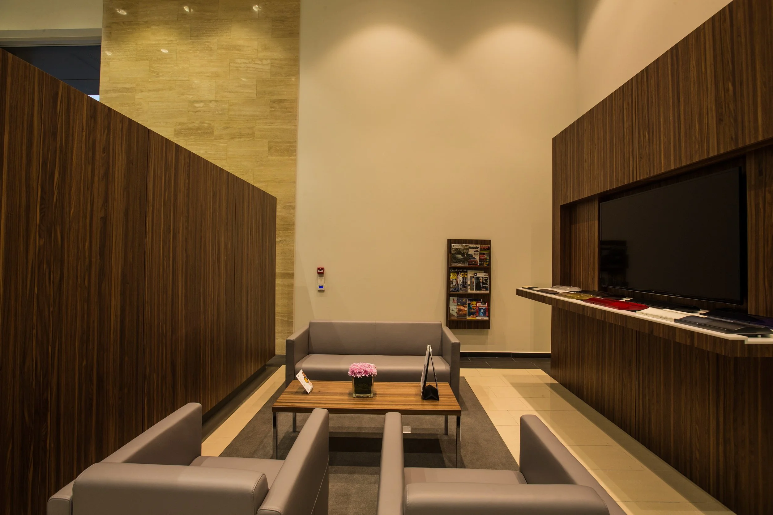 A modern waiting area with beige and wood-paneled walls, a gray sofa, a small wooden coffee table with a pink flower arrangement, magazines, and informational signage, illuminated by soft lighting.