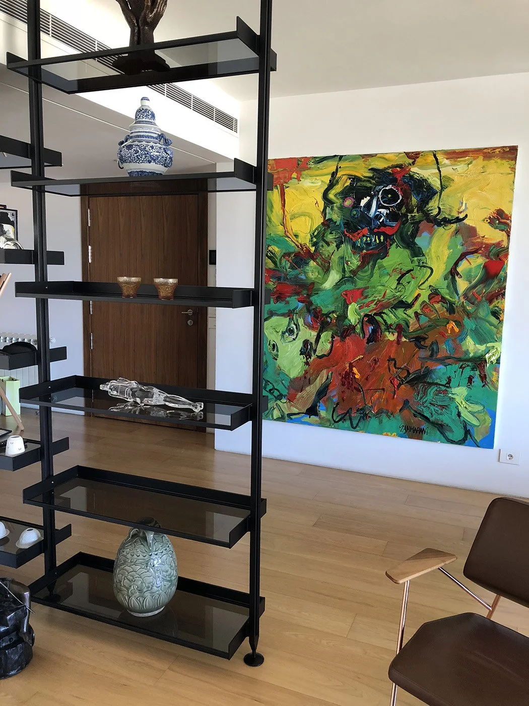 An interior space featuring a wooden door, a modern open black shelving unit with decorative items such as a blue and white porcelain jar, small bowls, and glass sculptures. A large, colorful abstract painting with vibrant green, red, yellow, and bla