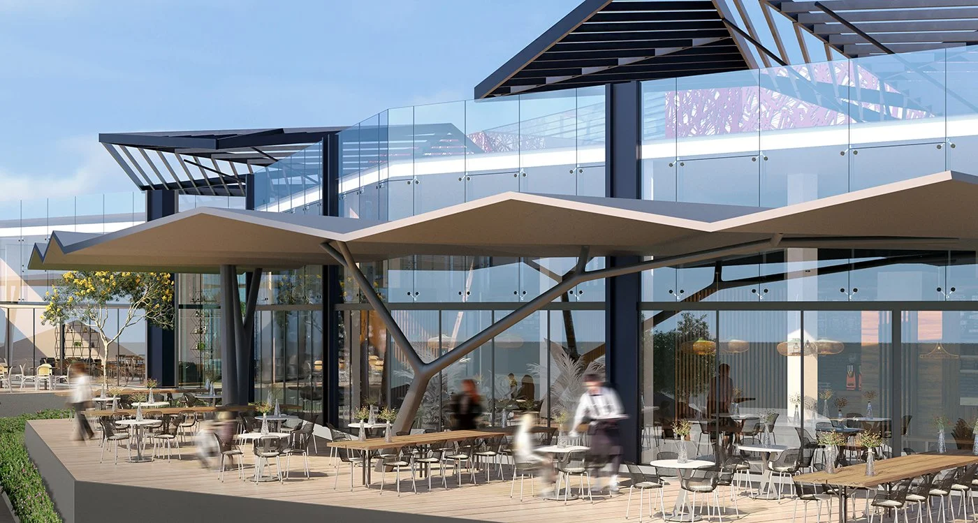 Modern building with glass walls and large outdoor patio with tables and chairs, featuring an canopy and architectural supports, blurred people walking, and trees in the background.
