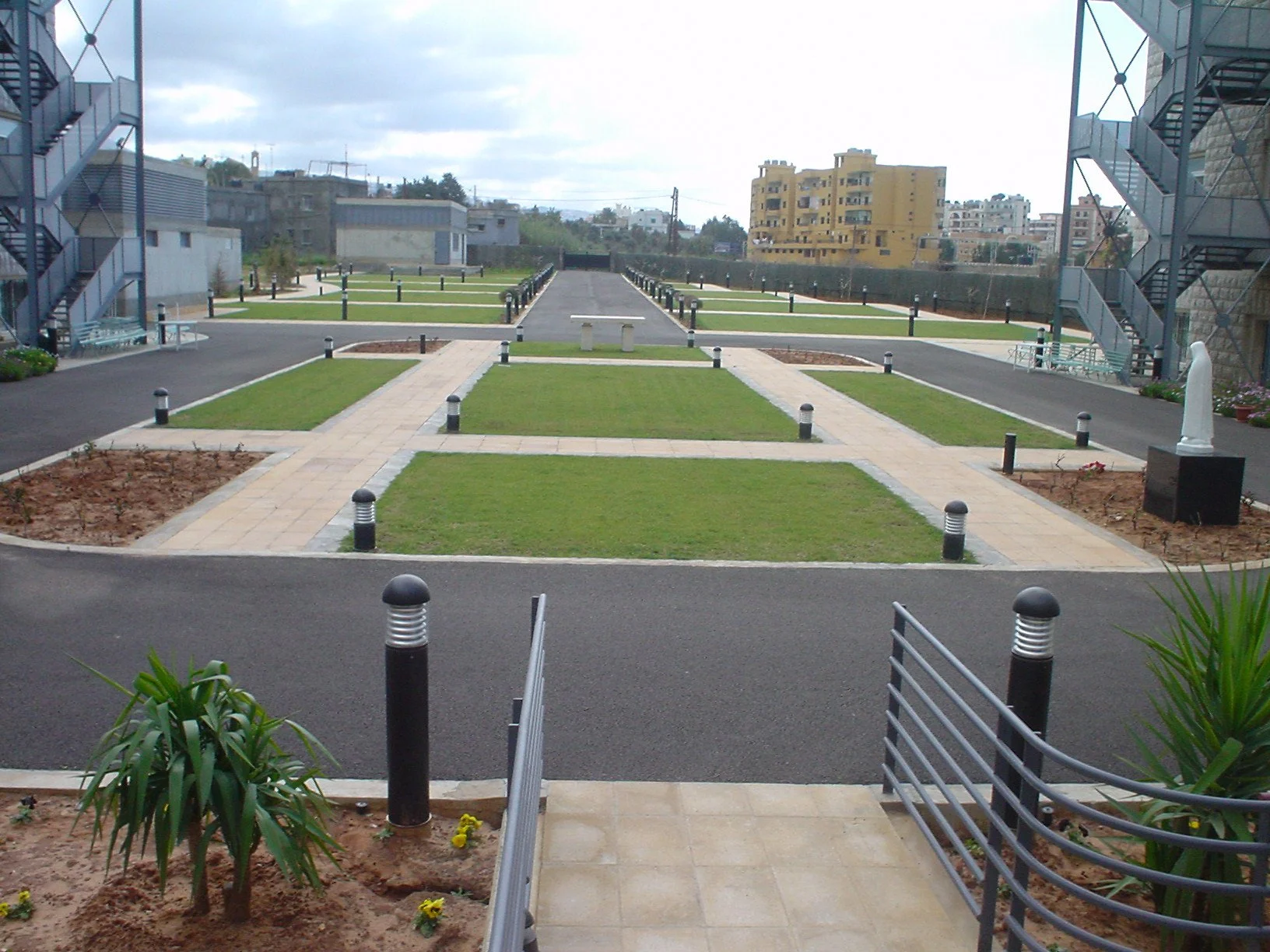 A modern outdoor space with paved walkways, green lawns, small flower beds, and decorative lighting fixtures, flanked by staircases on either side leading to an upper level, with a view of apartment buildings in the background, overcast sky.