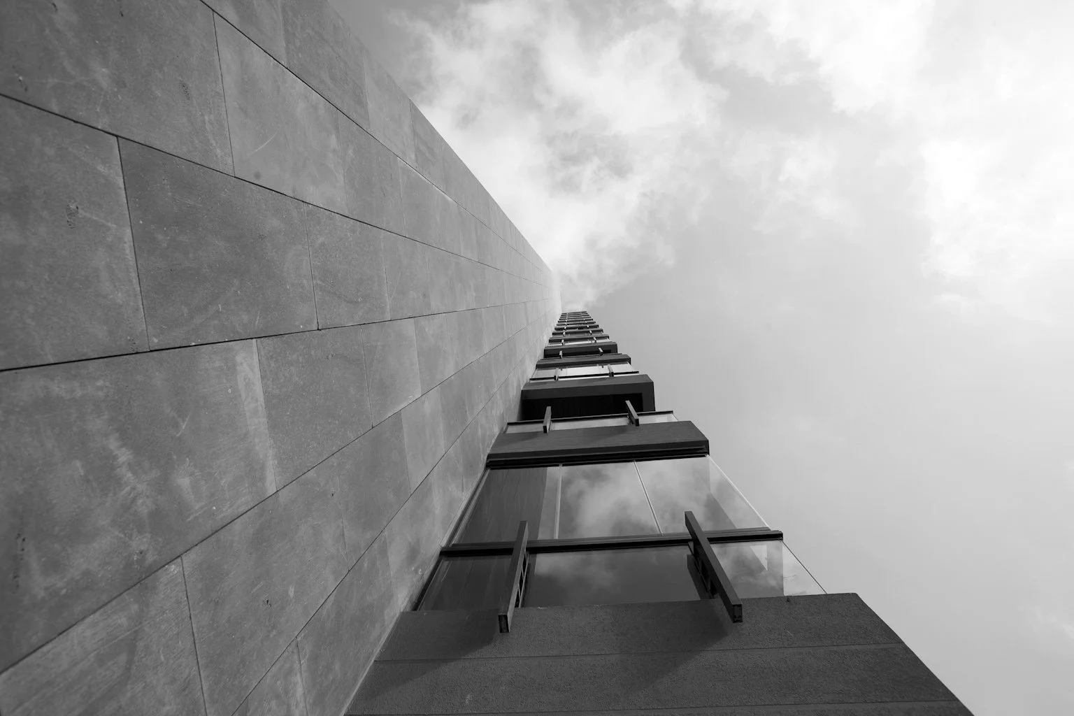 Black and white photo of a tall modern building taken from ground level looking upward, with a cloudy sky in the background.