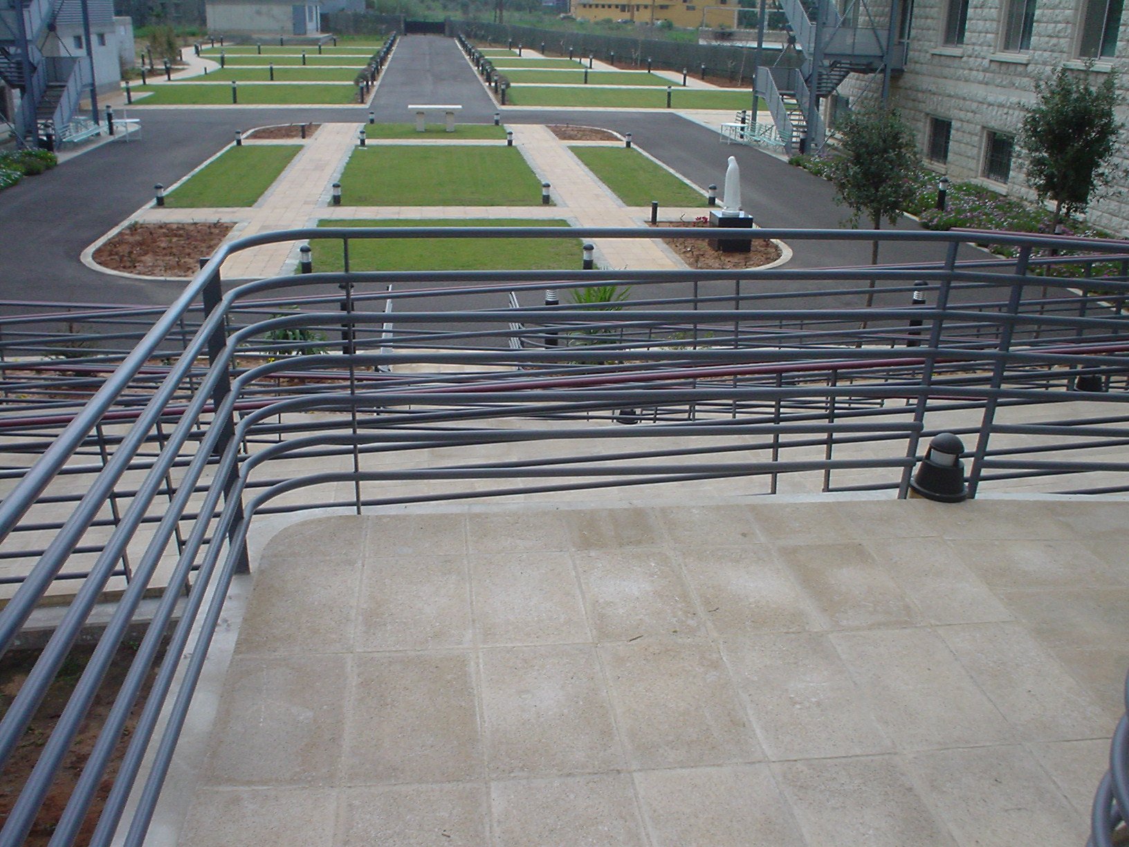 View from a concrete staircase with metal railings overlooking a courtyard with green grass, pathways, small trees, and surrounding buildings.