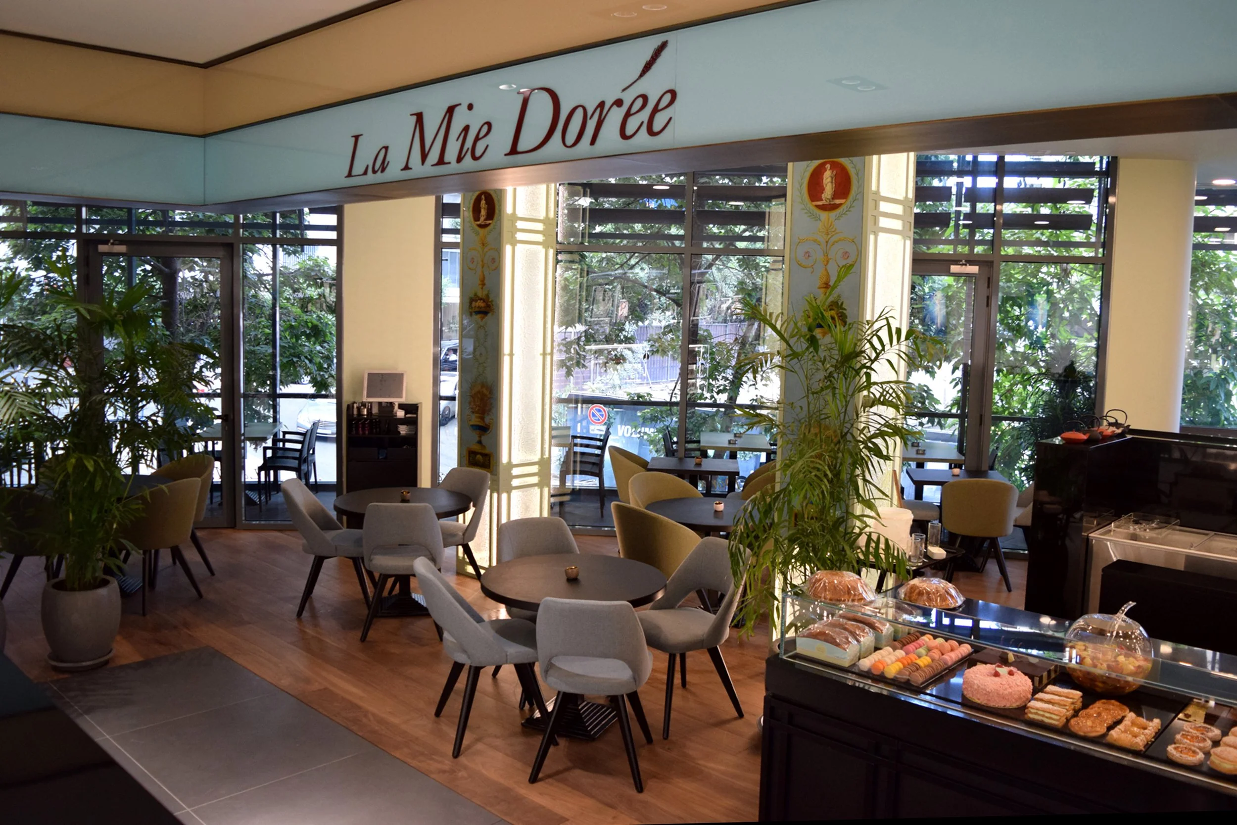Interior of a cafe named La Mie Dorée with tables, chairs, and a display of baked goods and desserts near large windows filled with greenery outside.