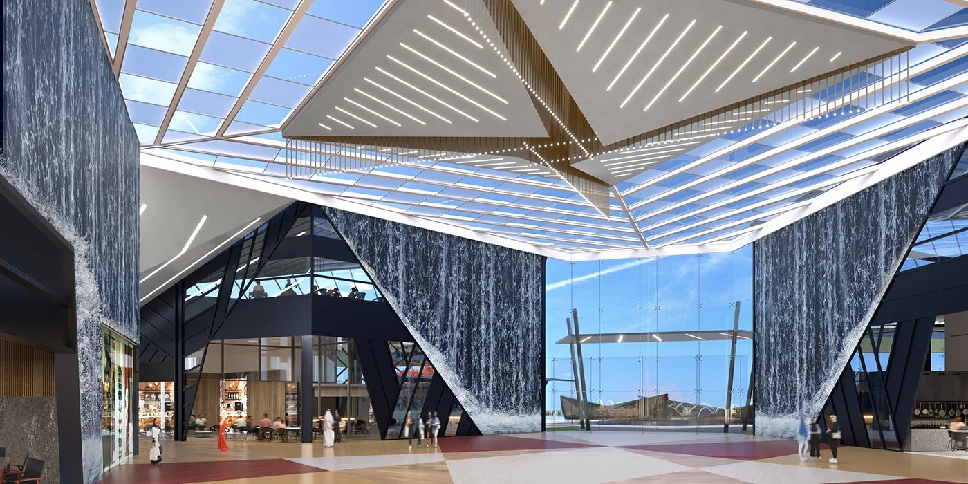 Interior of a modern building with a large glass ceiling, waterfall feature on the walls, and an open space with people walking and sitting.