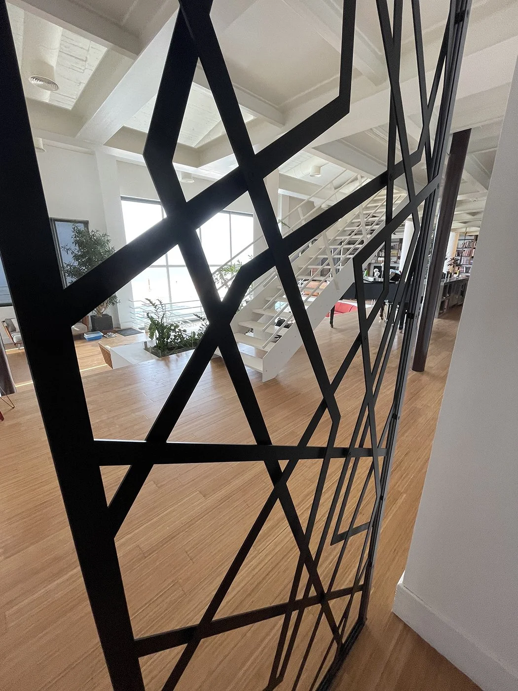 Interior view of a modern building with a white staircase, large windows, and wooden floors, seen through a decorative black metal partition with geometric patterns.