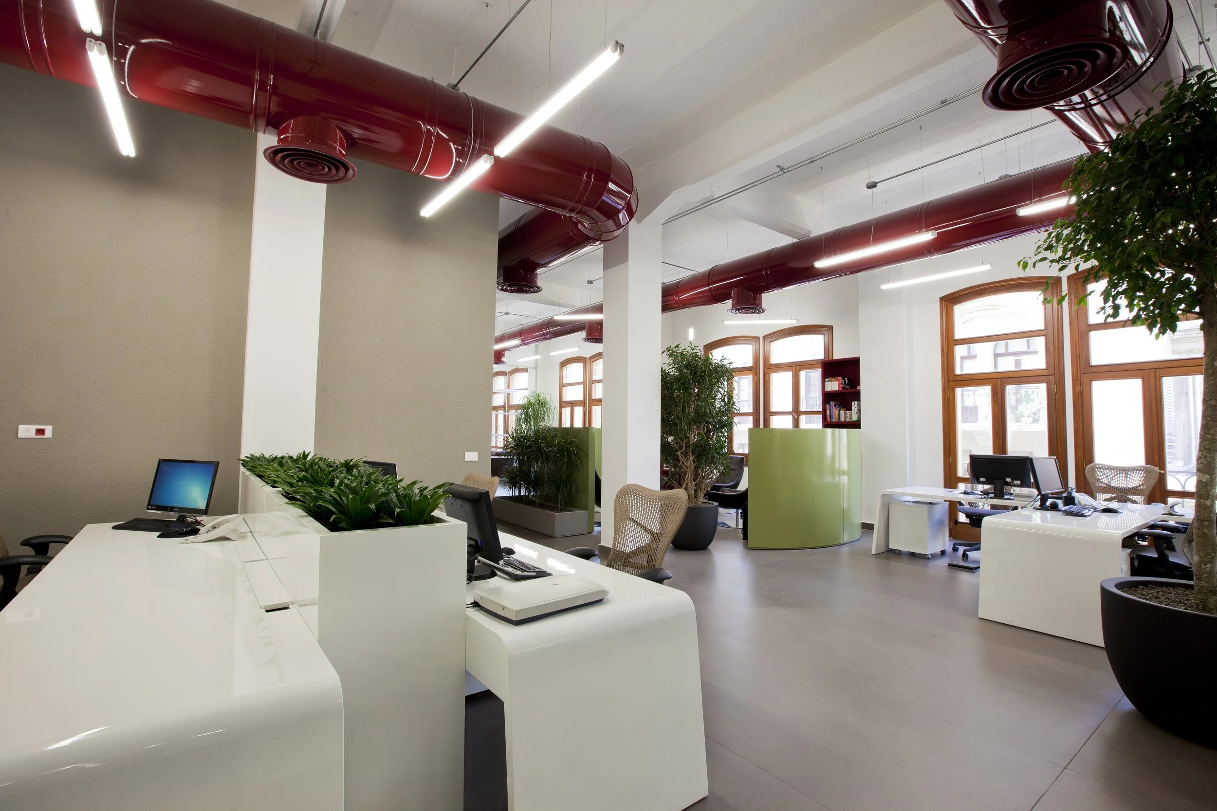 Modern office space with white desks, computer monitors, and office chairs, featuring large windows, potted plants, and red ceiling ducts.