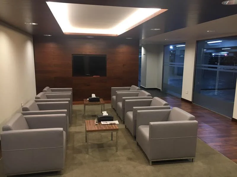 A modern waiting area with nine light gray armchairs arranged in three rows facing a large flat screen TV mounted on a dark wooden wall. There are two small wooden tables with glasses and tissue boxes in front of the chairs, and large windows on the 