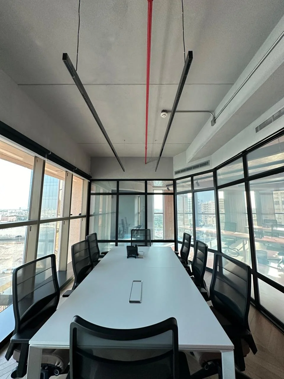 Modern conference room with a long white table, black mesh chairs, glass walls, daylight, city view, ceiling with black linear lighting fixtures, and a red ceiling pipe.