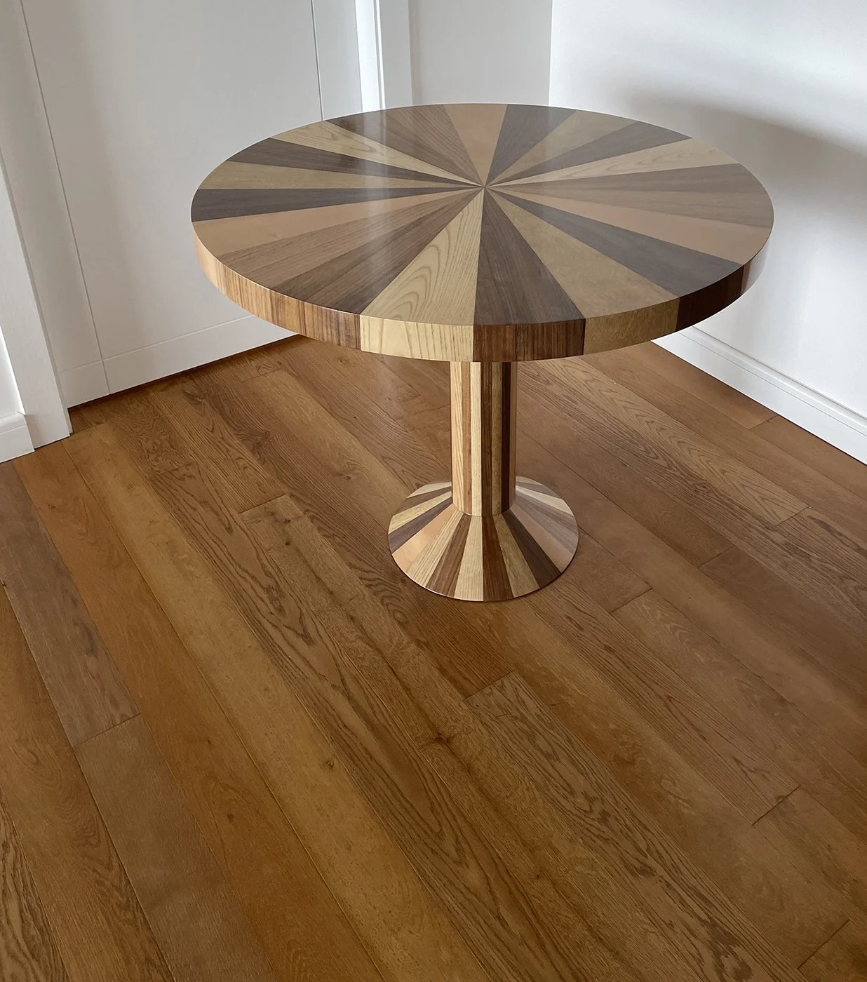 A round wooden table with a geometric pattern on the top, supported by a single pedestal base with a matching pattern, standing on a wooden floor.
