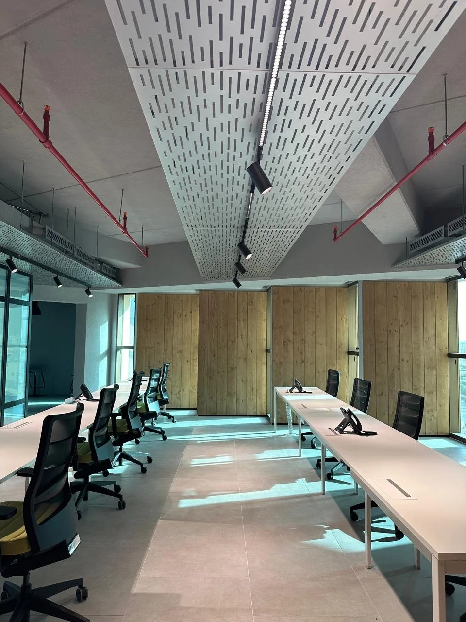 Empty modern conference room with black office chairs at white desks with black phones, large windows with natural light, wood panel walls, and a perforated ceiling with track lighting.