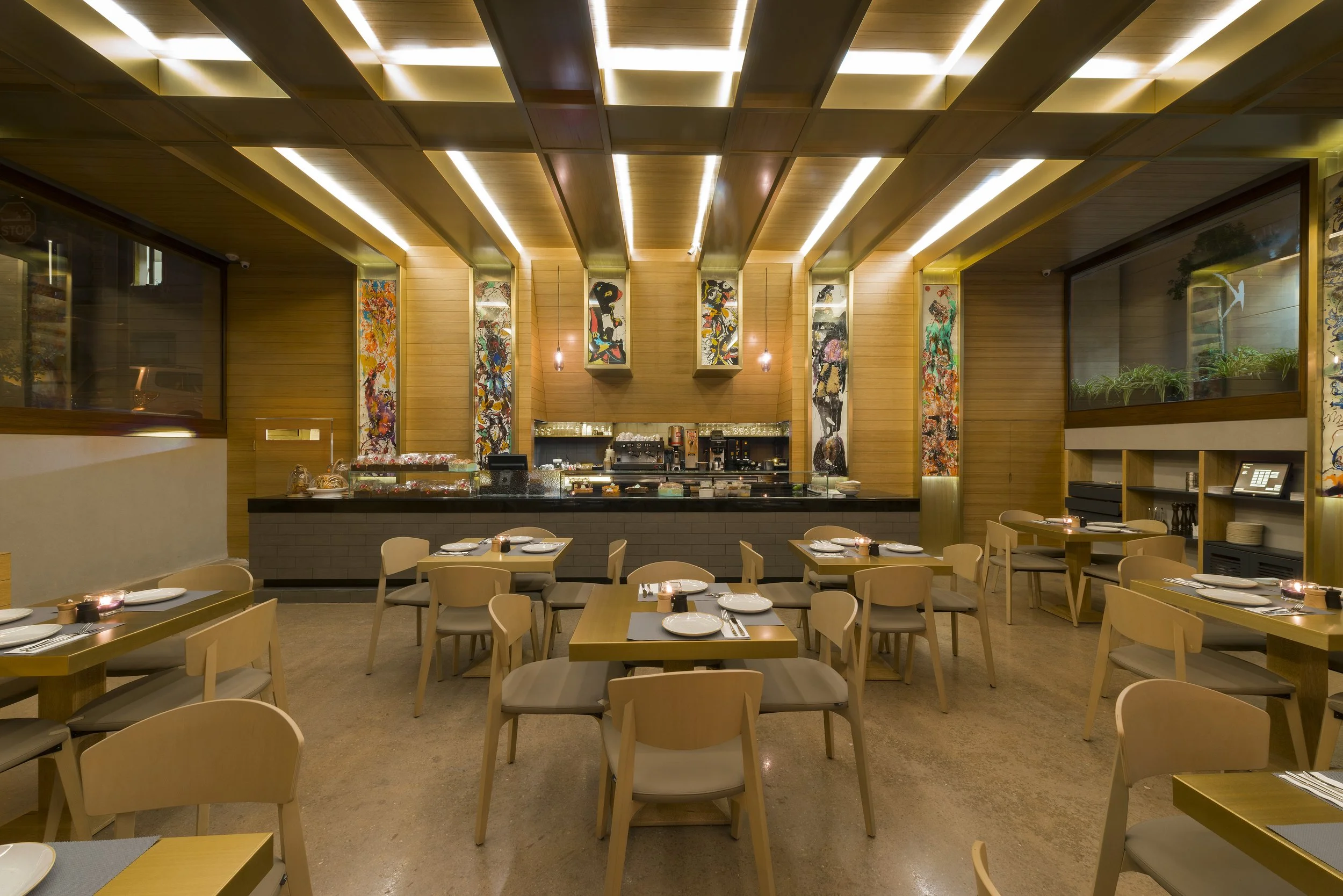Interior of a modern restaurant with wooden walls and ceiling, featuring colorful abstract artwork, tables set with plates and napkins, and a counter area with coffee machines and shelves.