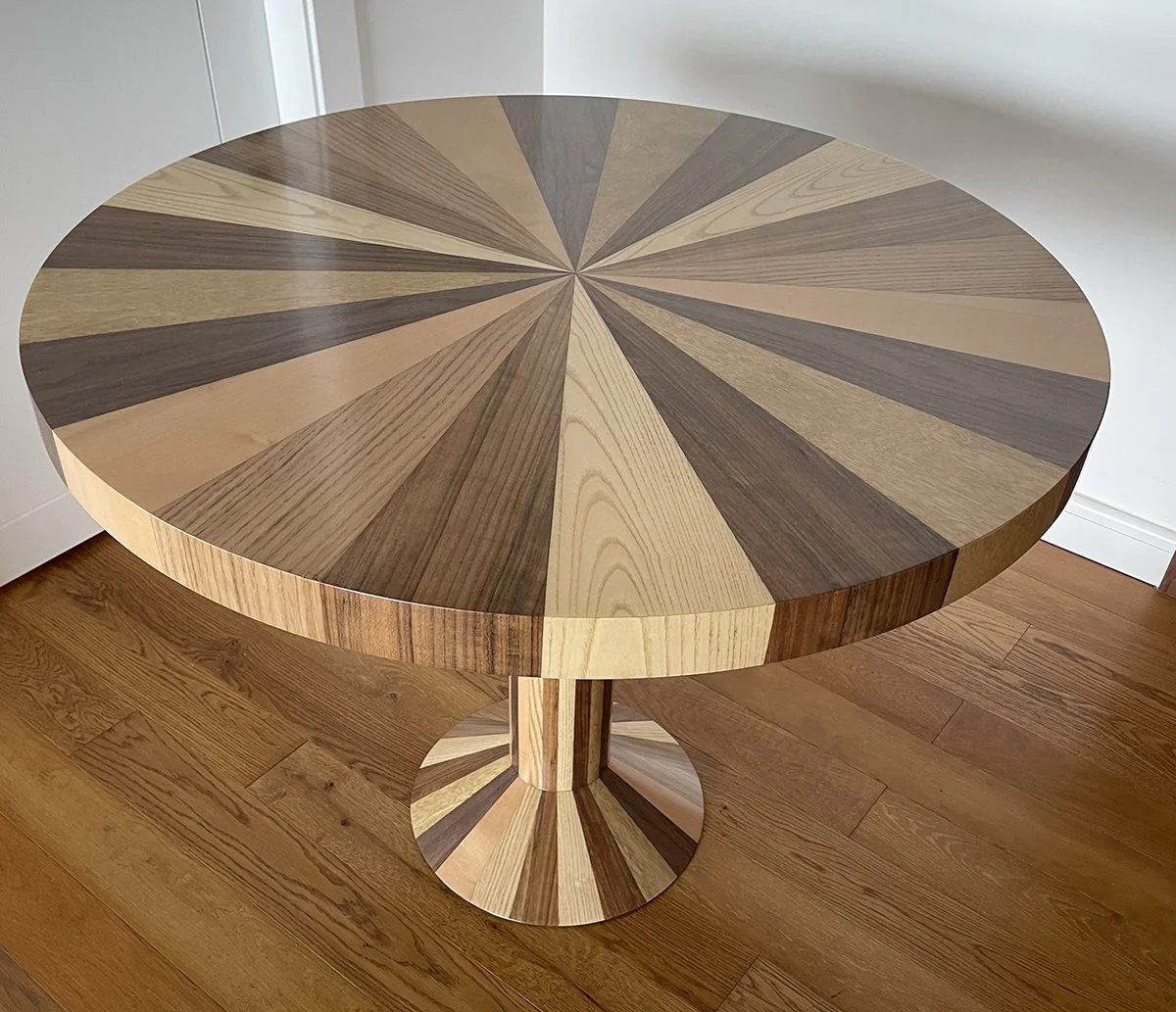Round wooden table with a multi-toned wood pattern top, standing on a matching striped base, placed on a hardwood floor near a white wall.