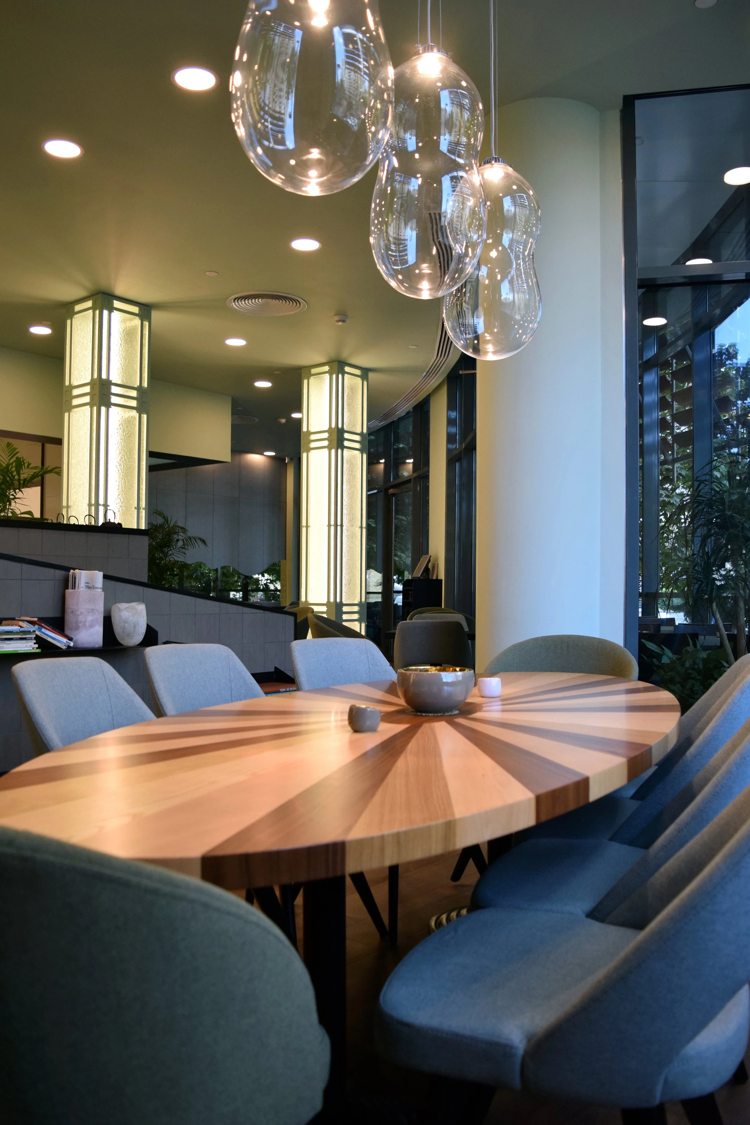 A modern restaurant interior with a round wooden table surrounded by light gray upholstered chairs, decorative glass pendant lights hanging from the ceiling, and large windows with a view of trees outside.