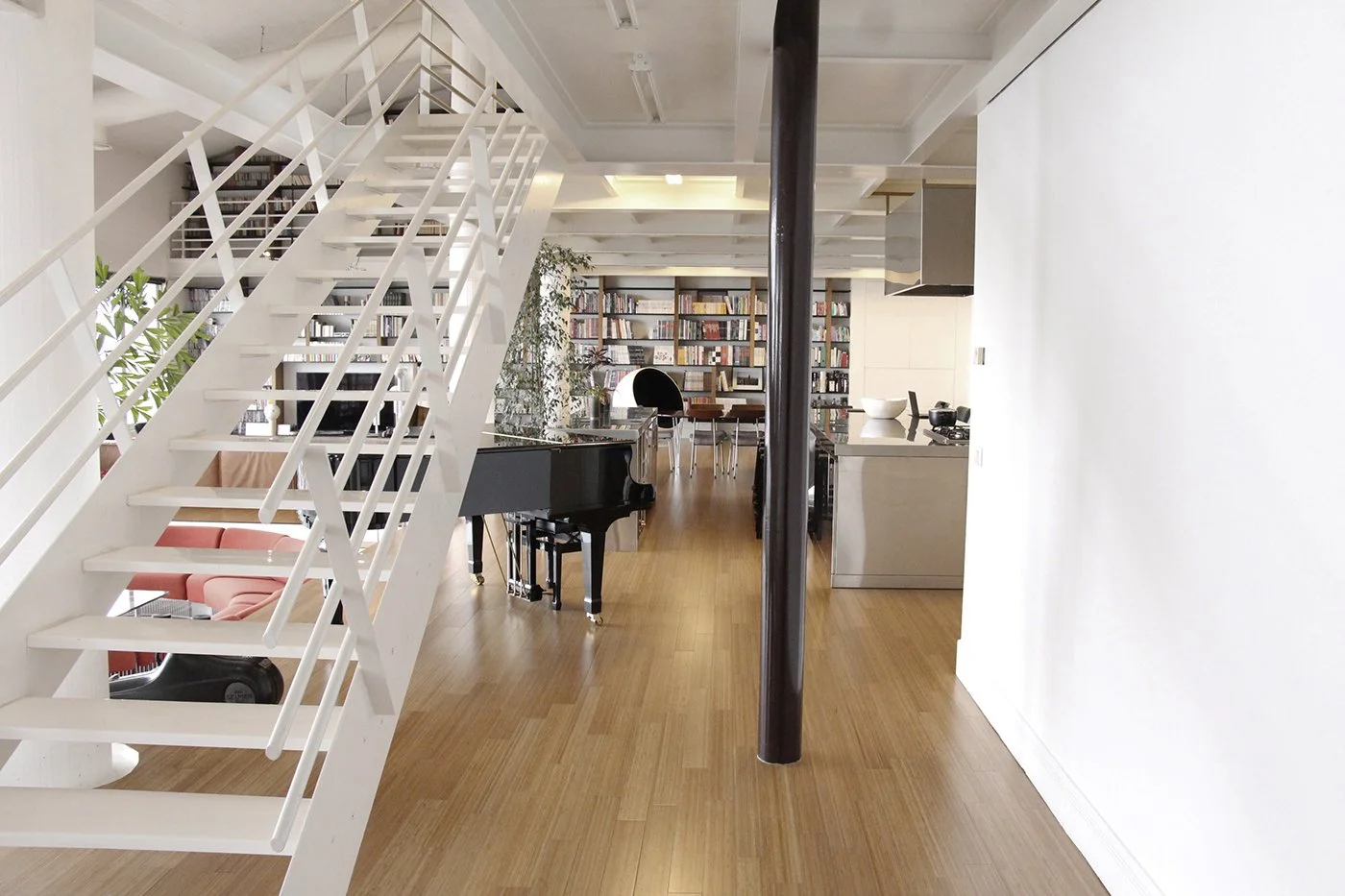 Interior of a modern apartment with a white staircase, black grand piano, wooden floor, and a bookshelf wall in the background.