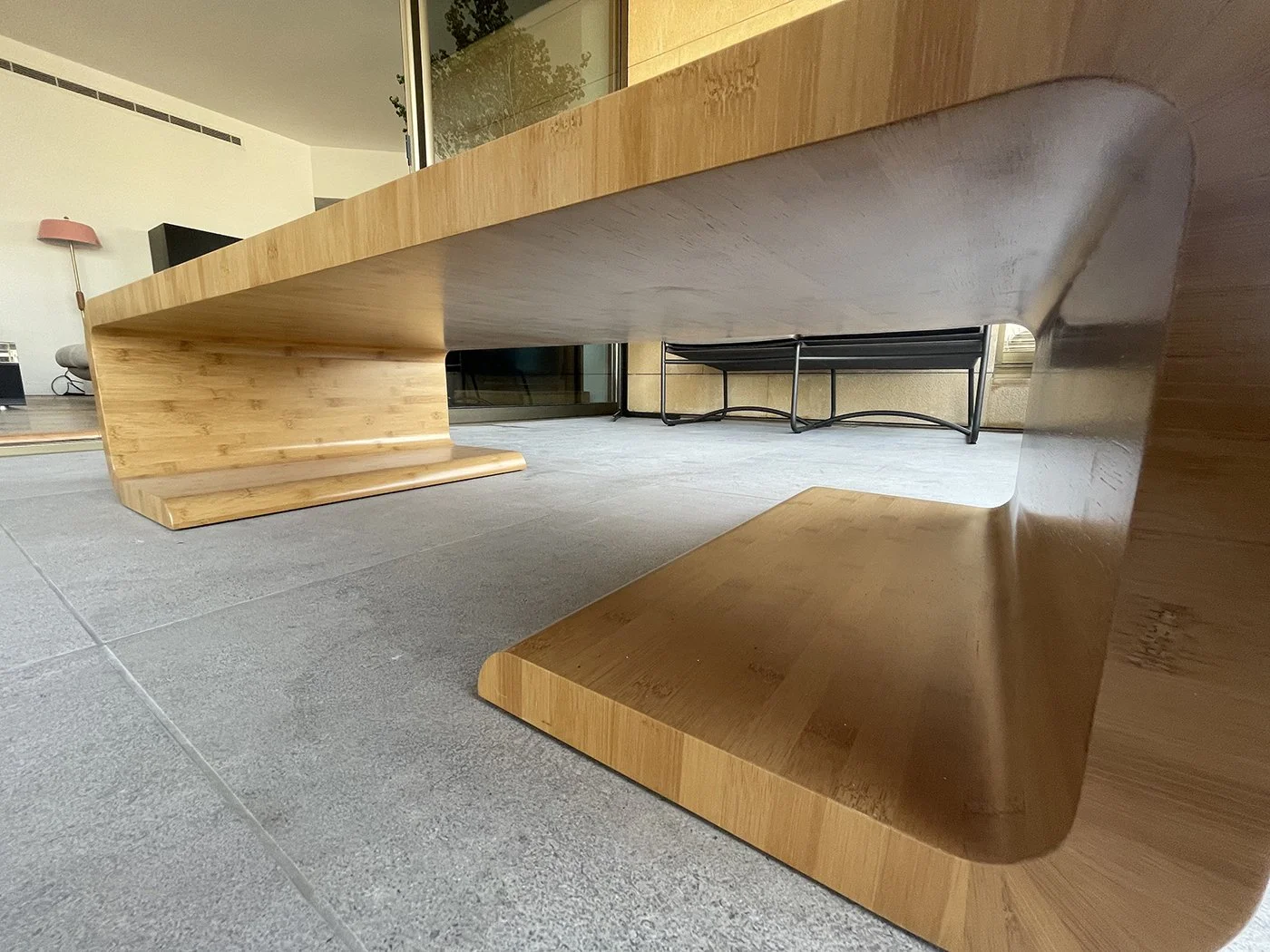 Low-angle view of a modern wooden coffee table with a unique curved design, situated on a tiled indoor space near large glass doors.