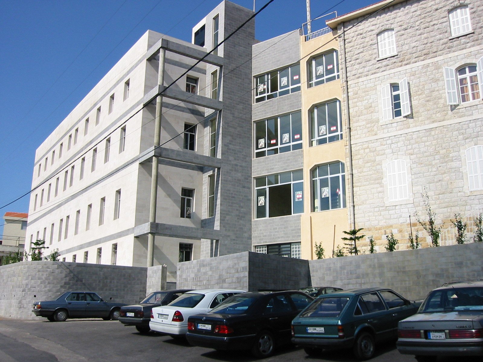 Modern multi-story building with exposed grey concrete and glass windows, parked cars in the foreground, and a clear blue sky.