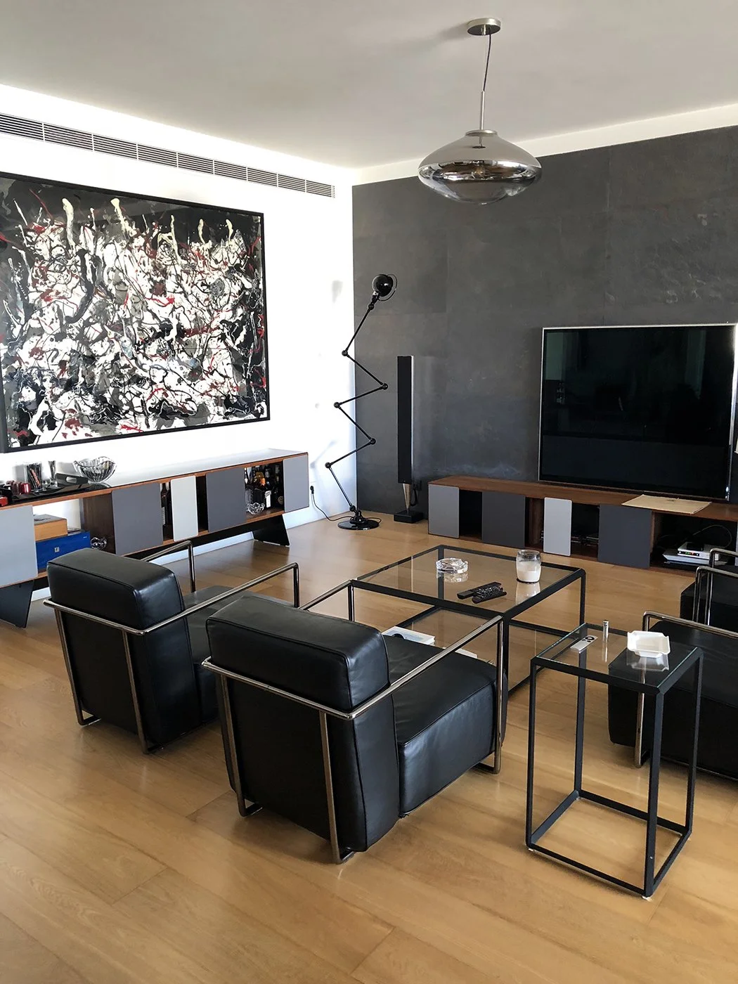 Modern living room with black leather chairs, wooden flooring, abstract art, a large TV, and contemporary lighting fixtures.