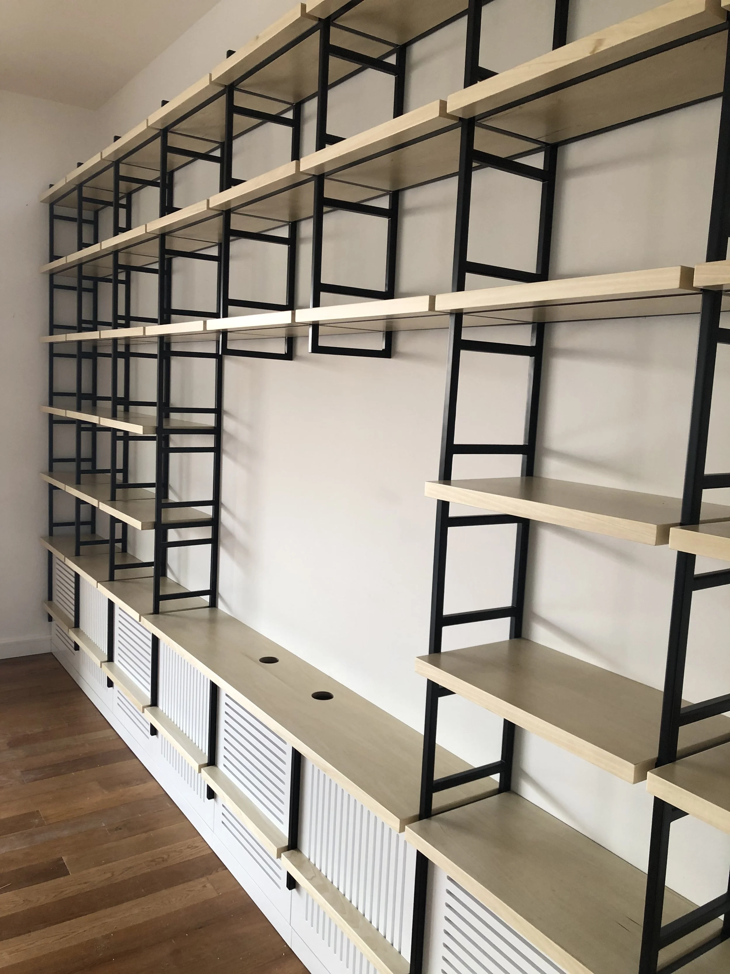 Empty modular shelving unit with light wooden shelves, black metal supports, and white vented cabinet storage at the bottom, set against a white wall and near a wooden floor.