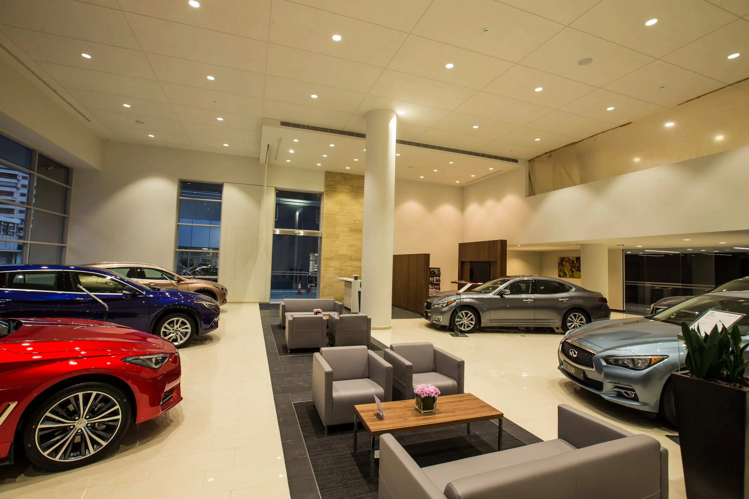 Interior of a modern car dealership with multiple luxury cars on display, including a red car in the foreground and various other cars like gray and brown, with a seating area and large windows showing buildings outside.