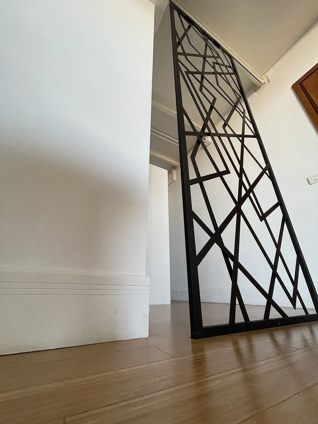 Interior of a room with a black metal decorative room divider with an abstract geometric pattern, wooden flooring, white walls, and a wooden cabinet partially visible on the right side.