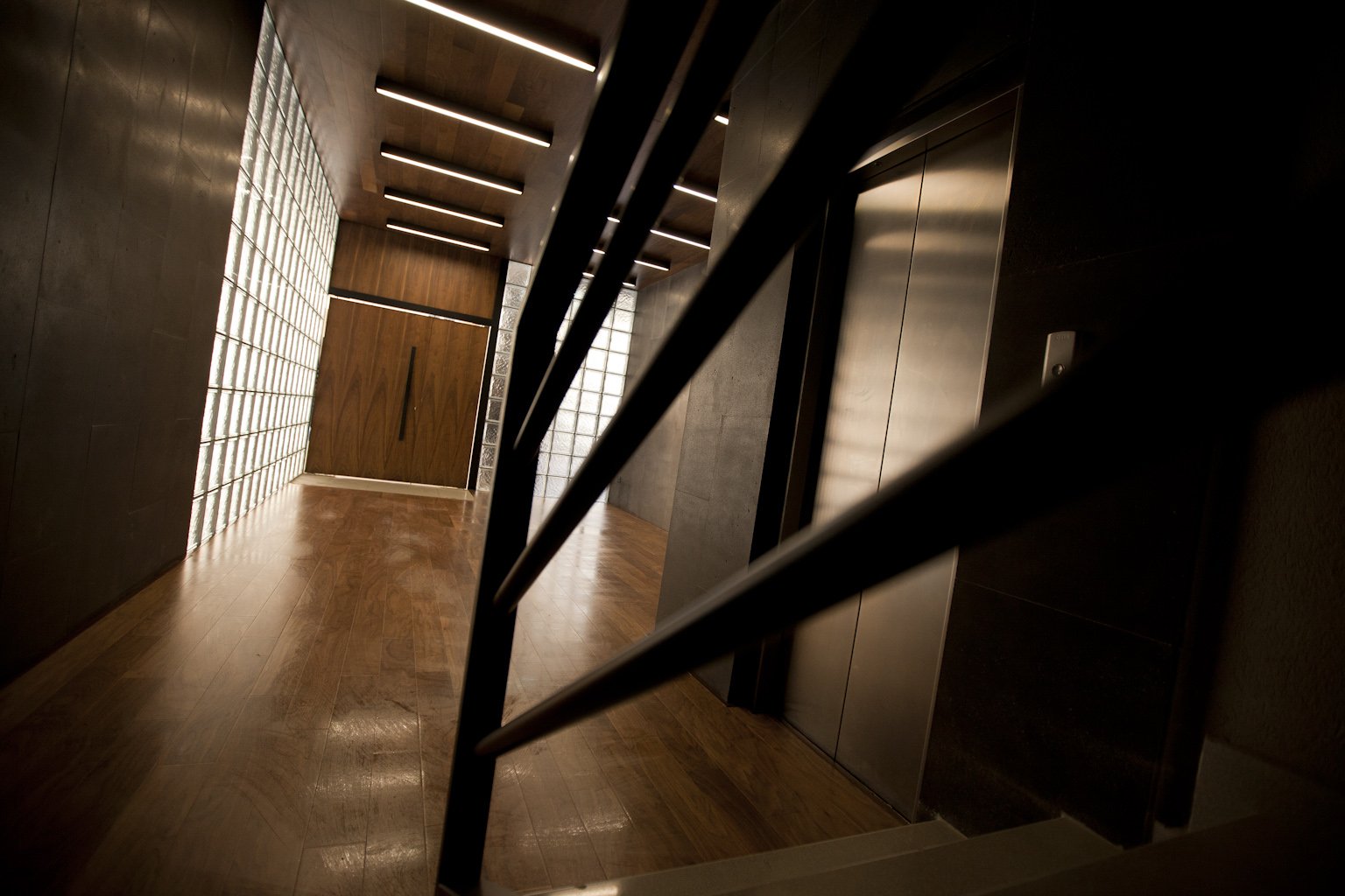 Interior view of a modern hallway with wooden floors, glass block windows on left, an elevator door on right, and a wooden door at the end.